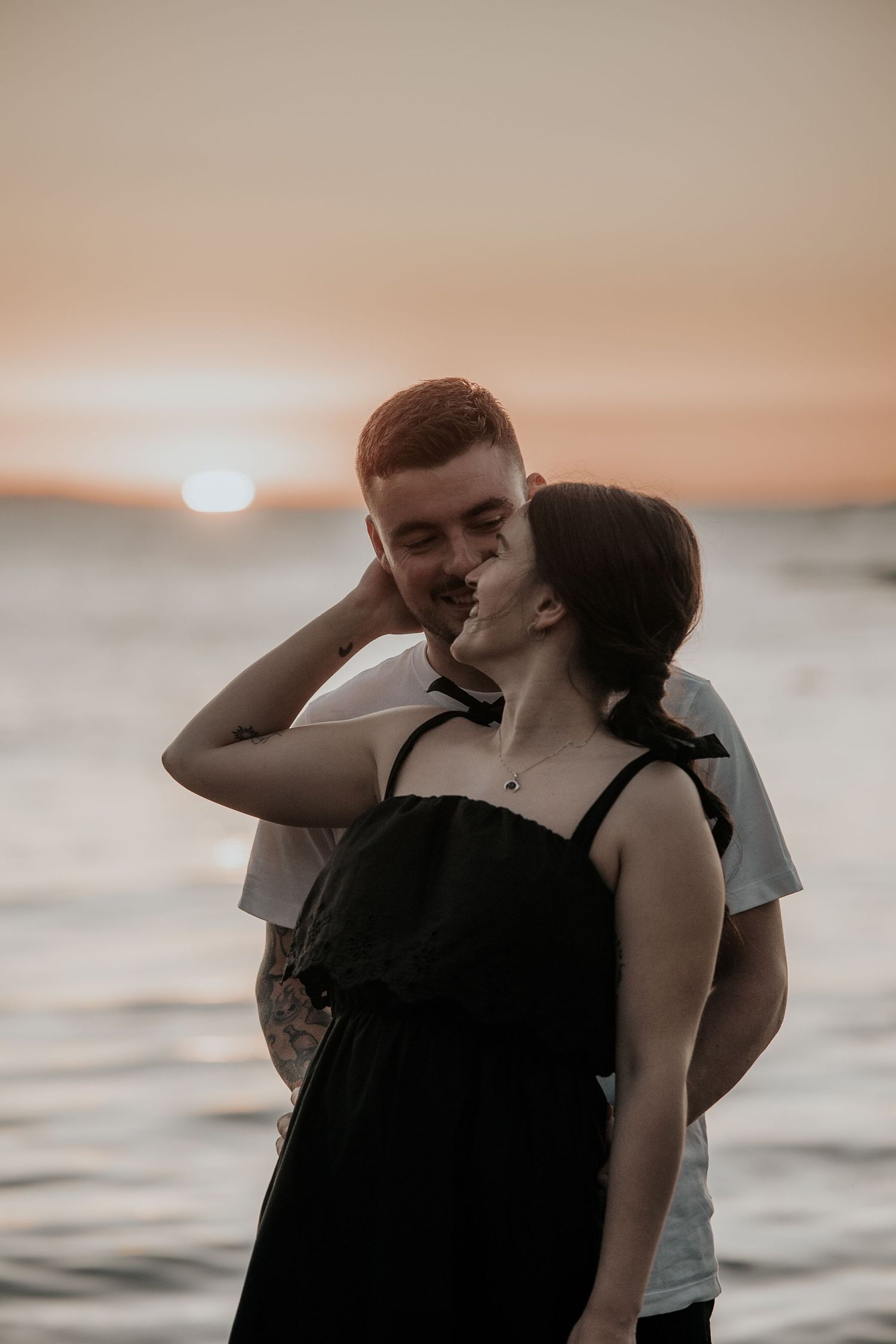 A romantic couple embraces during sunset at the beach with warm orange tones in the sky and ocean behind them.