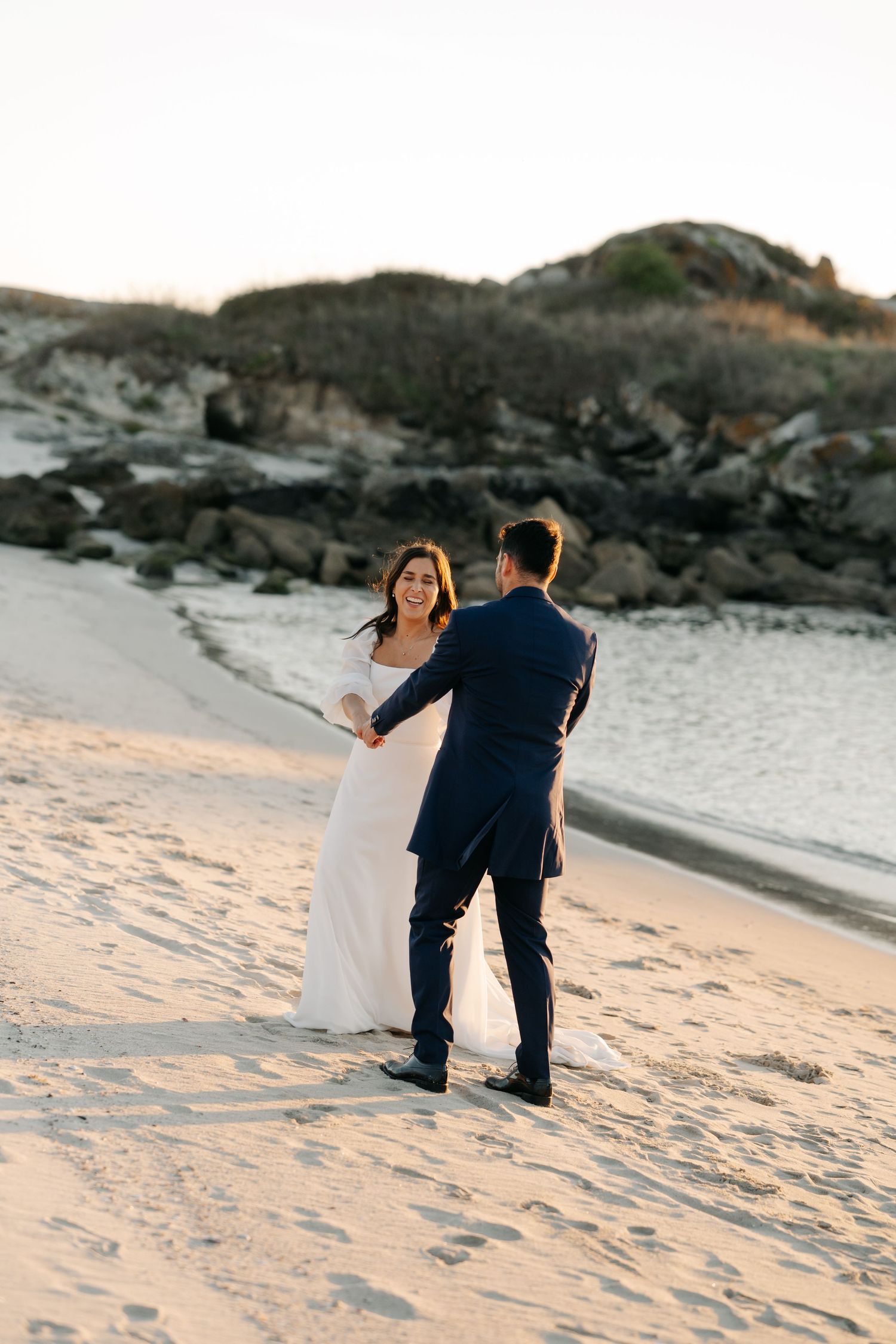Couple walking together on a sandy beach at sunset with rocky cliffs in the background.