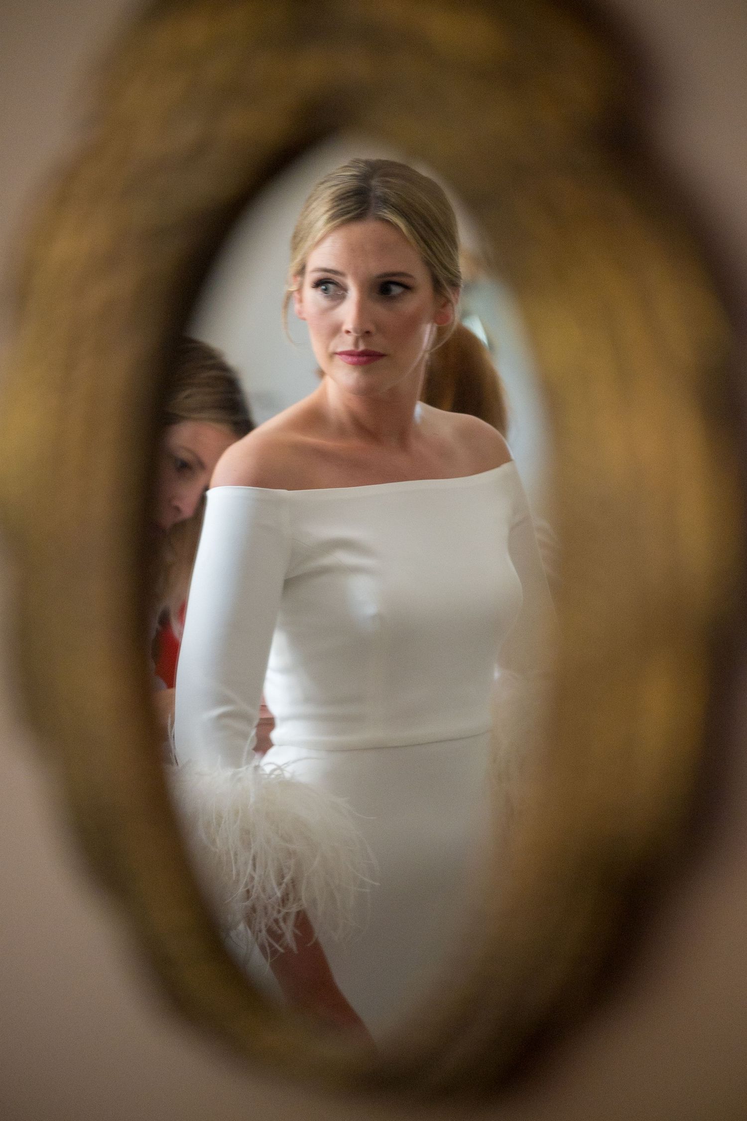 A bride in an elegant off-shoulder white dress with feather details seen through a circular frame.