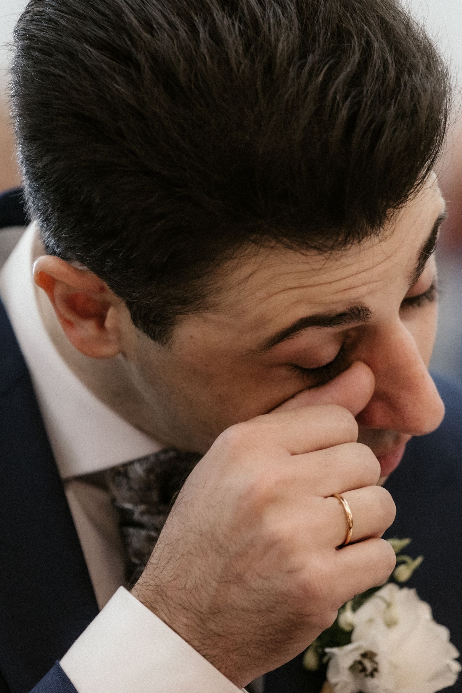 An emotional groom in a navy suit wipes tears from his eyes during a wedding ceremony.