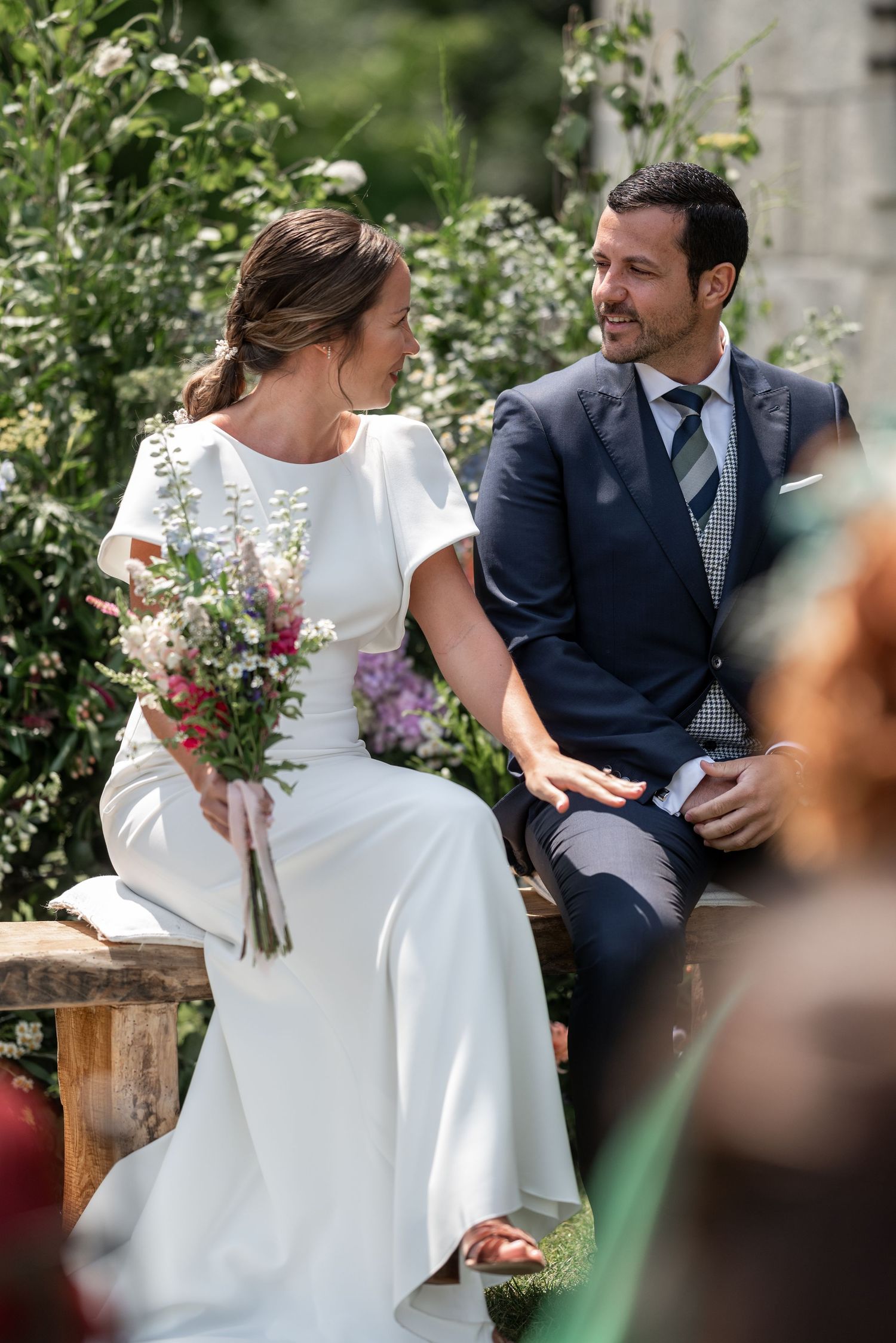 A couple sits on a bench in a garden setting with the bride holding a bouquet of wildflowers in her white dress.