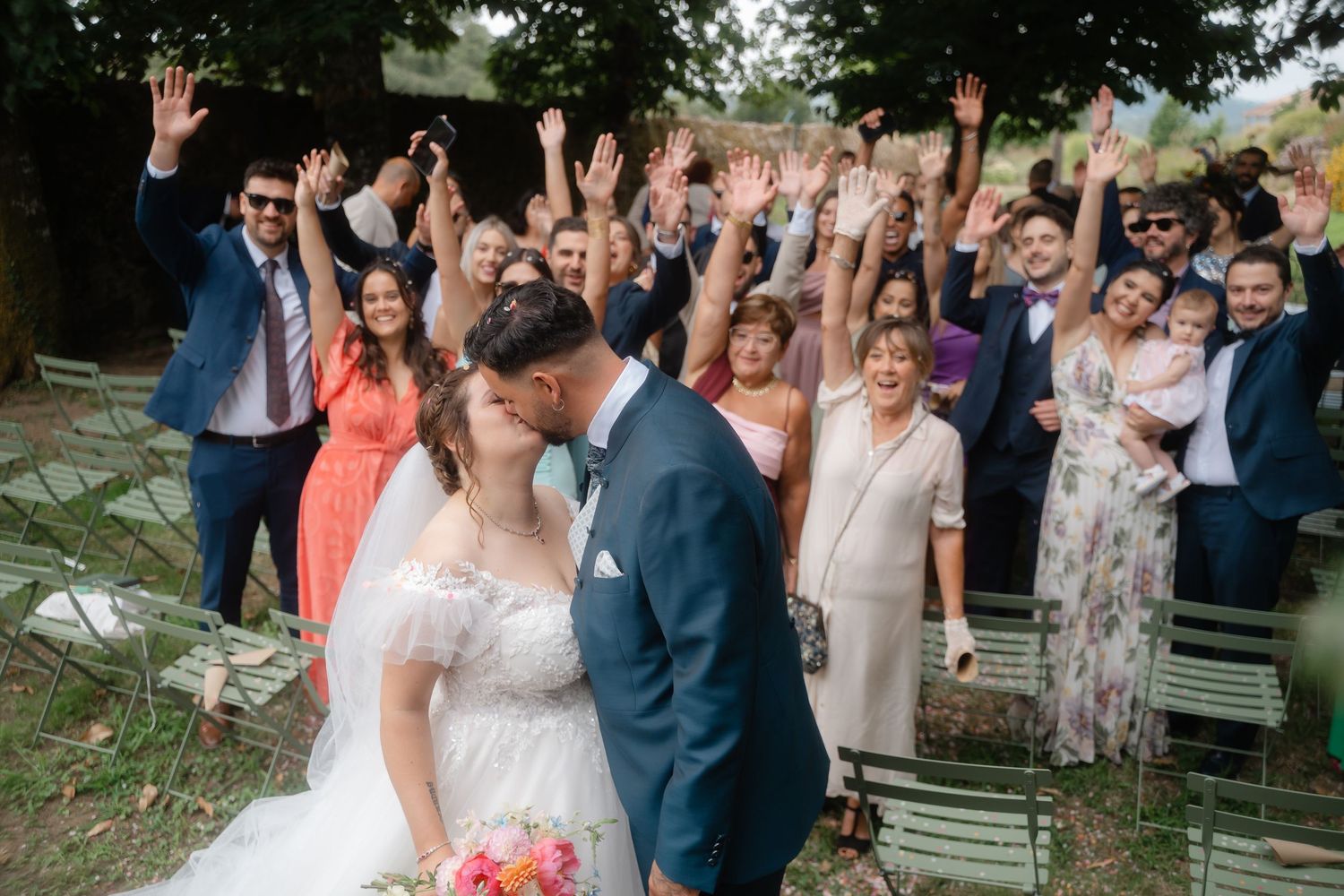 Wedding party celebrates outdoors with joyful poses and raised arms during summer celebration at garden venue.