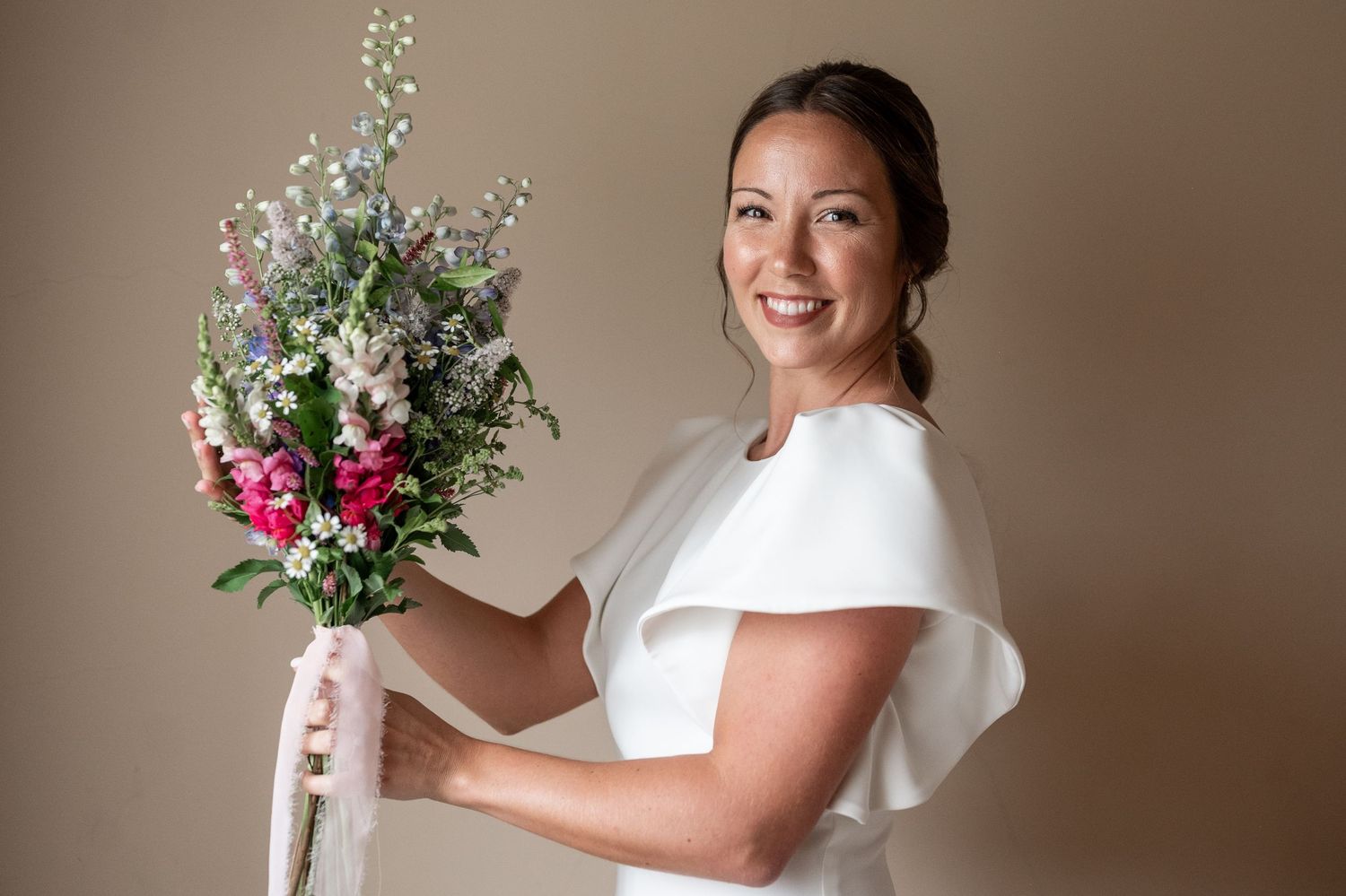 A person in a white wedding dress holds a beautiful bouquet of pink and white flowers against a neutral background.