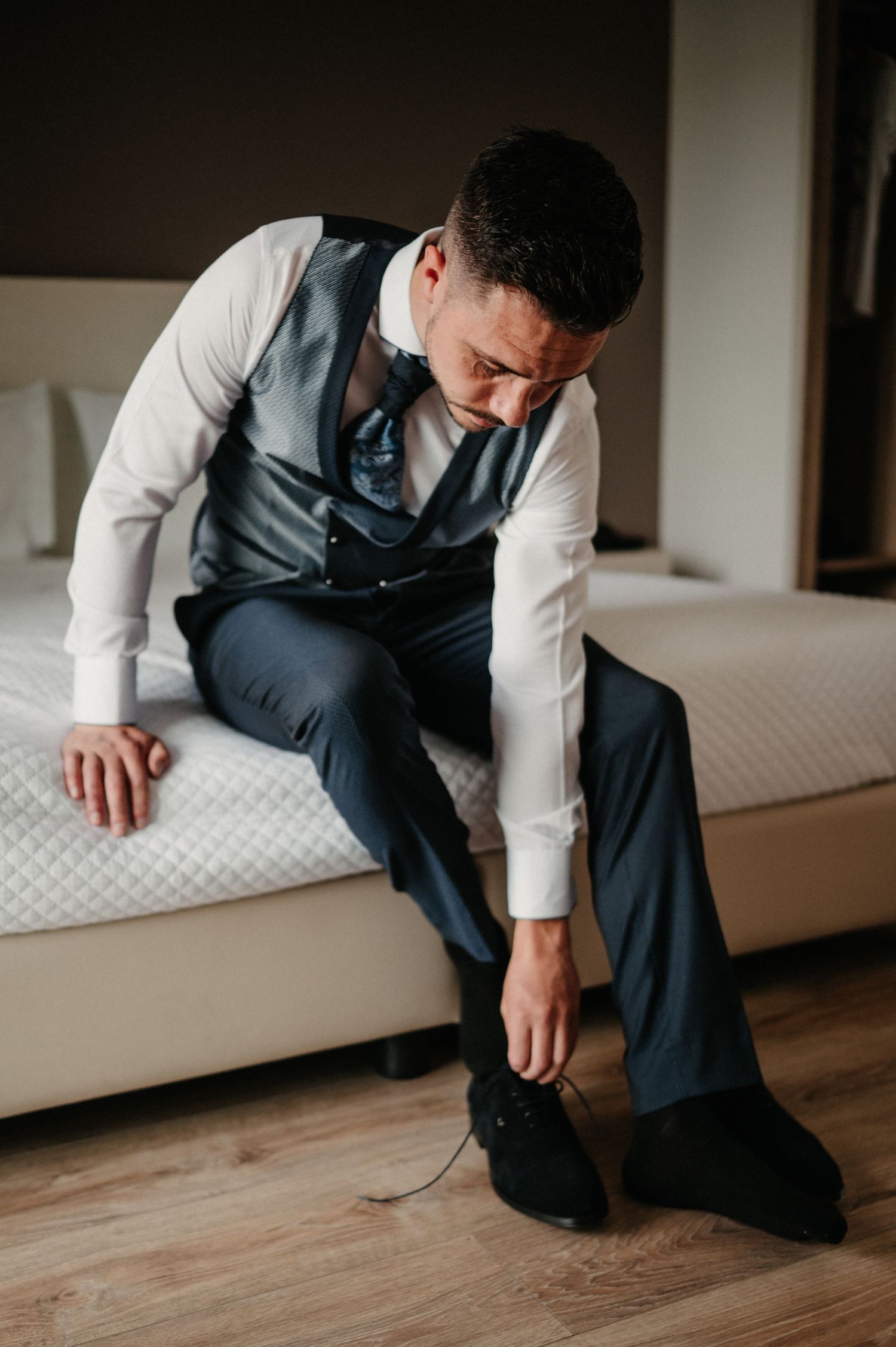 A person in formal attire sits on a bed while putting on black dress shoes for a special occasion.