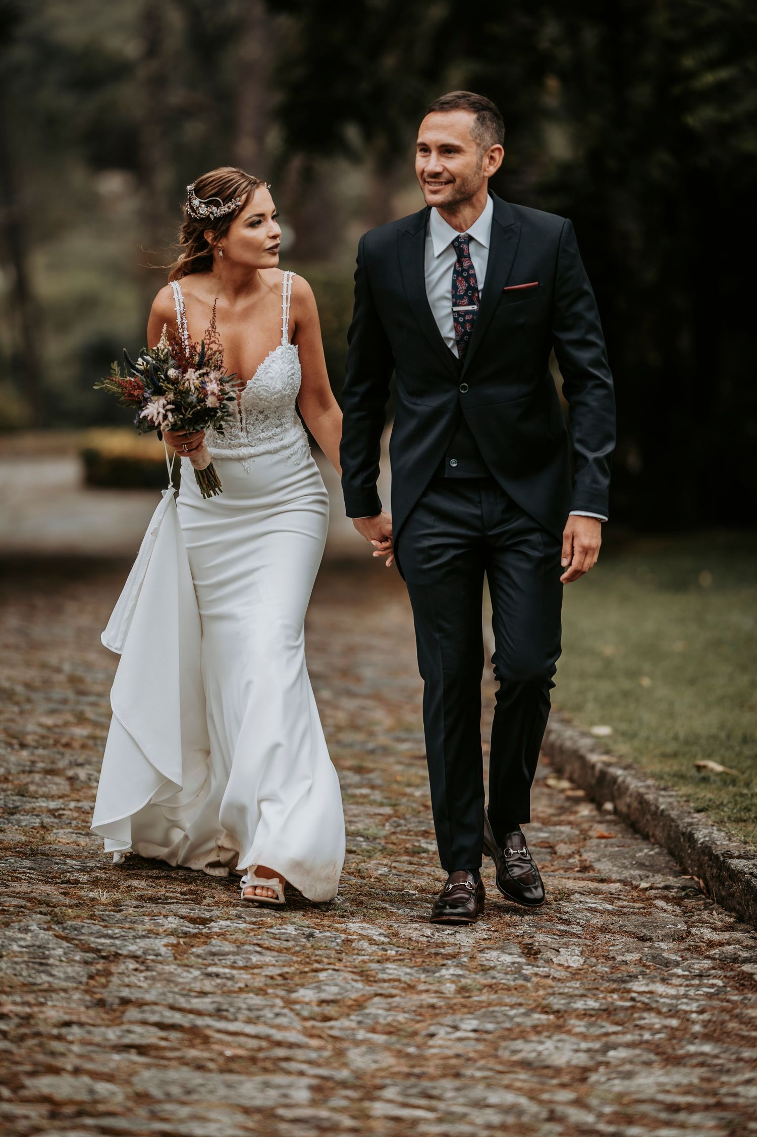 Bride in white dress and groom in suit walking together on stone path through autumn leaves.