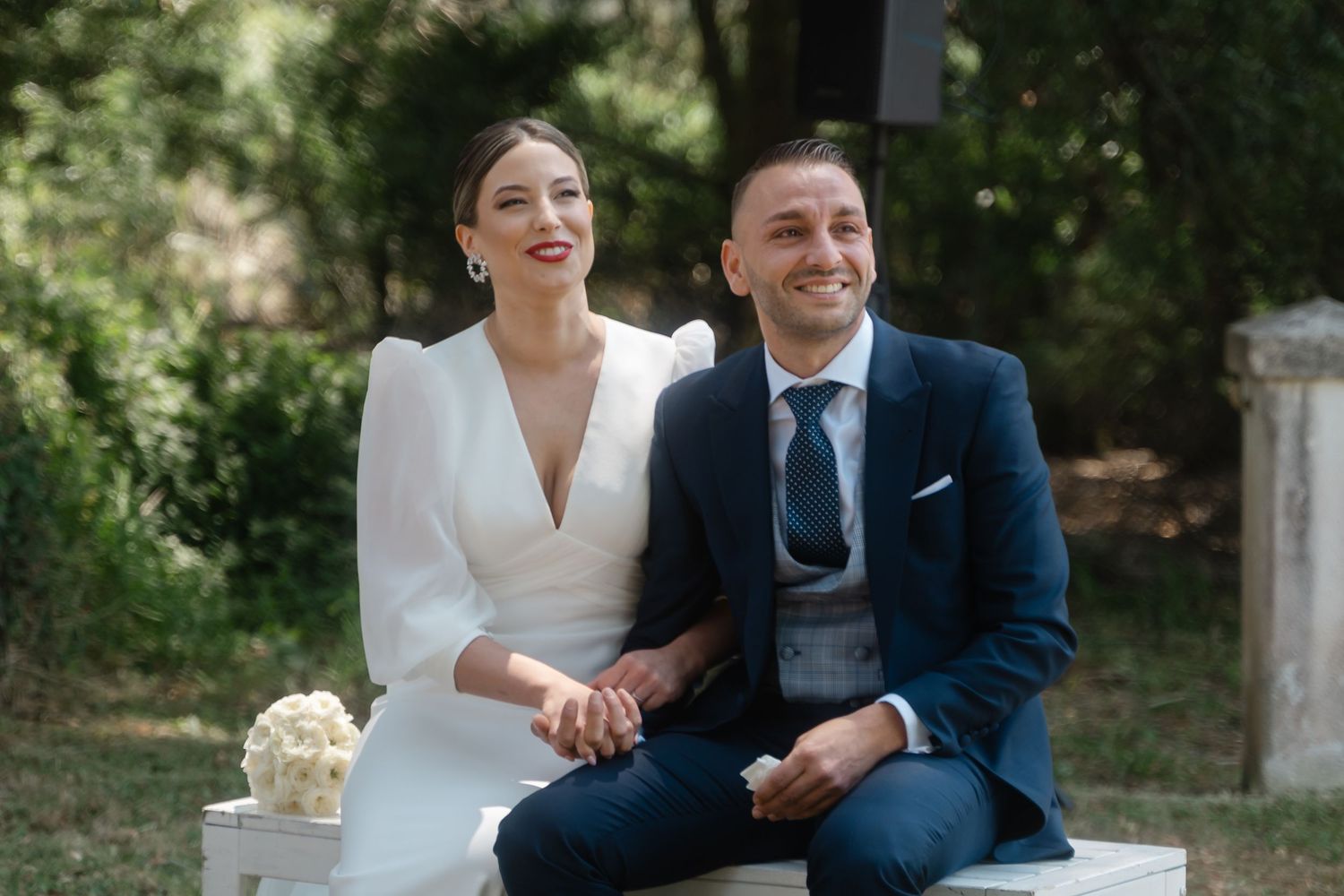 A couple in wedding attire sits together outdoors sharing a joyful moment in a garden setting.