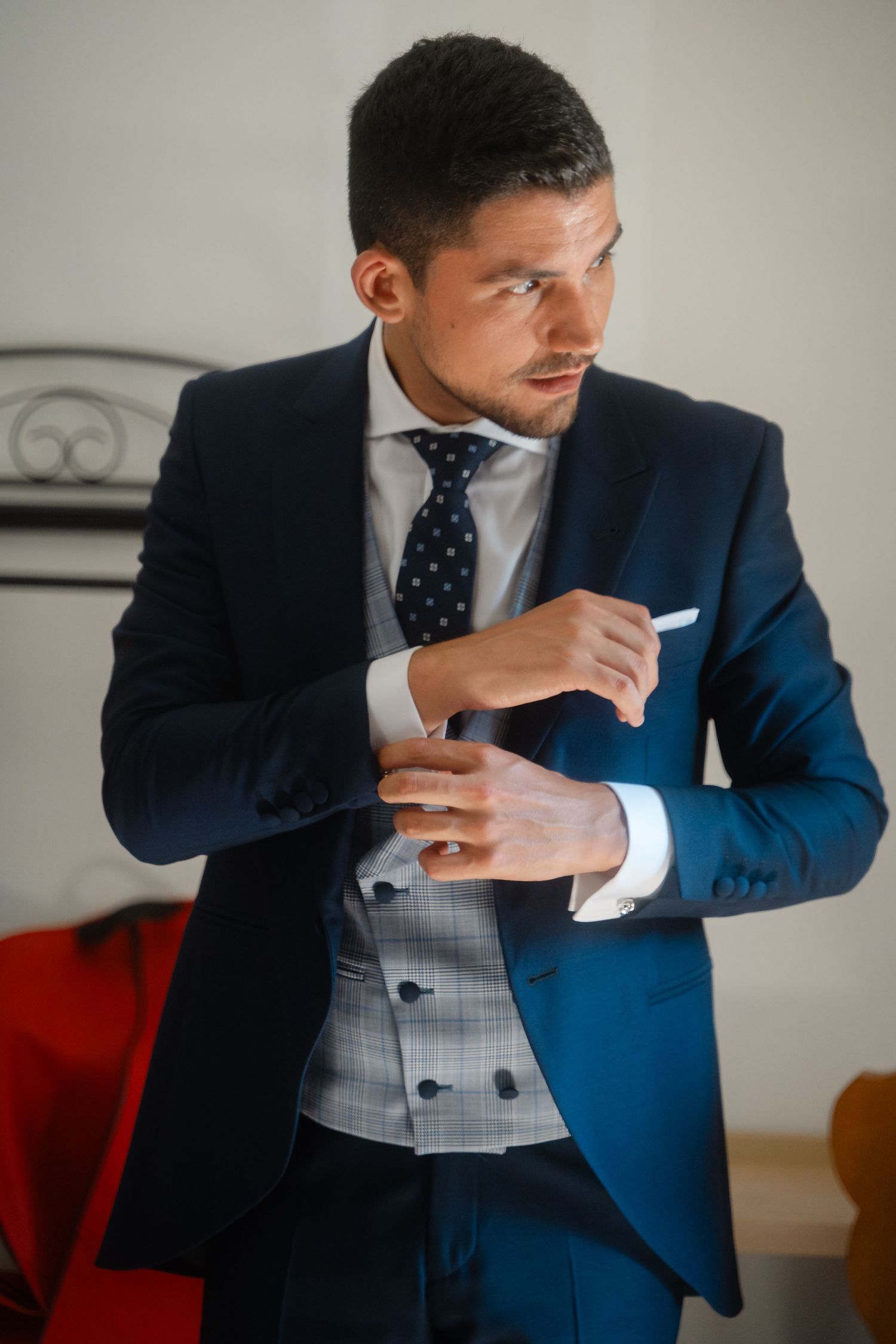 A person in a navy suit and polka dot tie adjusts their cuff while wearing a light gray waistcoat.