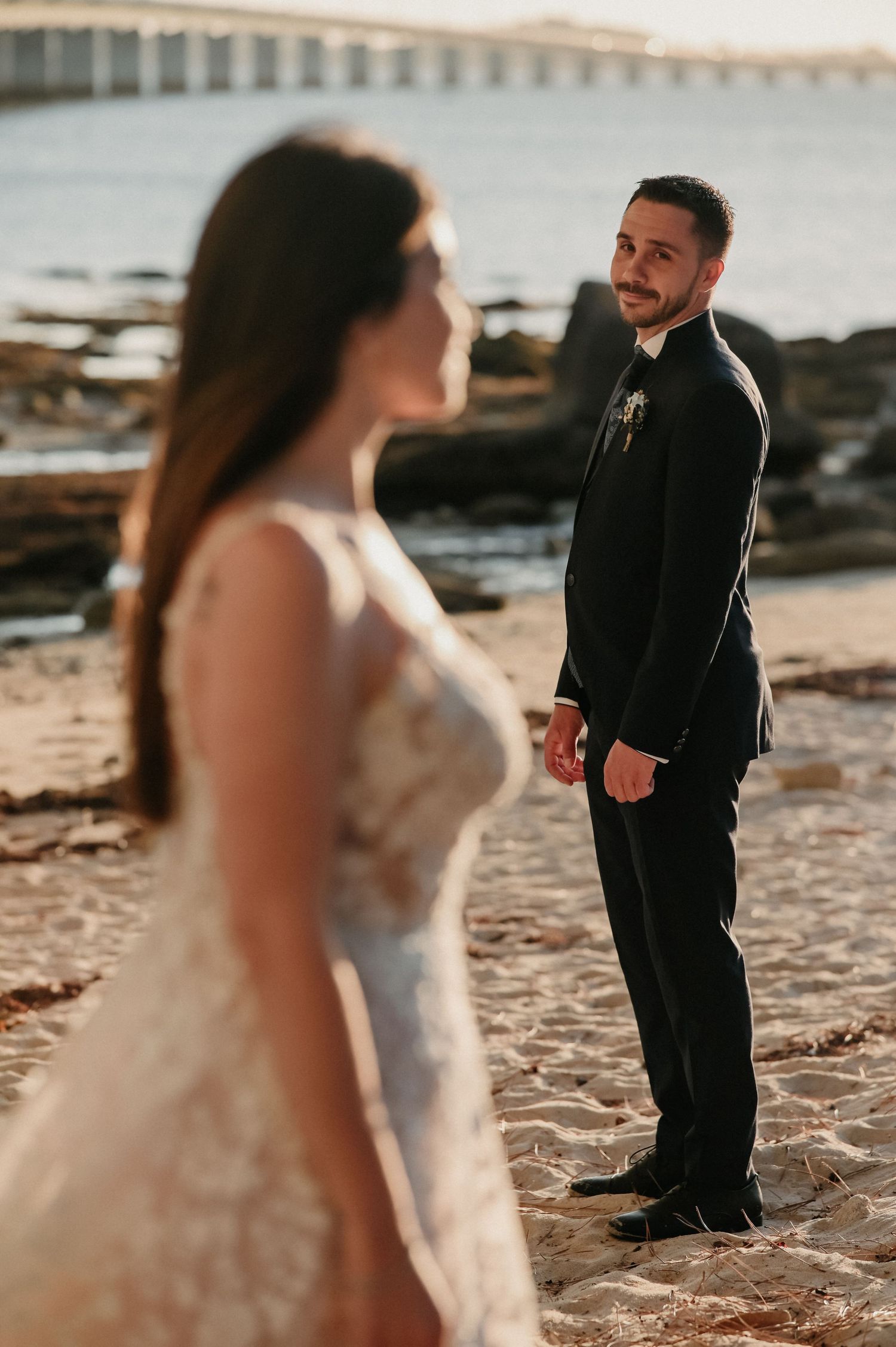 A couple in wedding attire poses on a beach during golden hour.