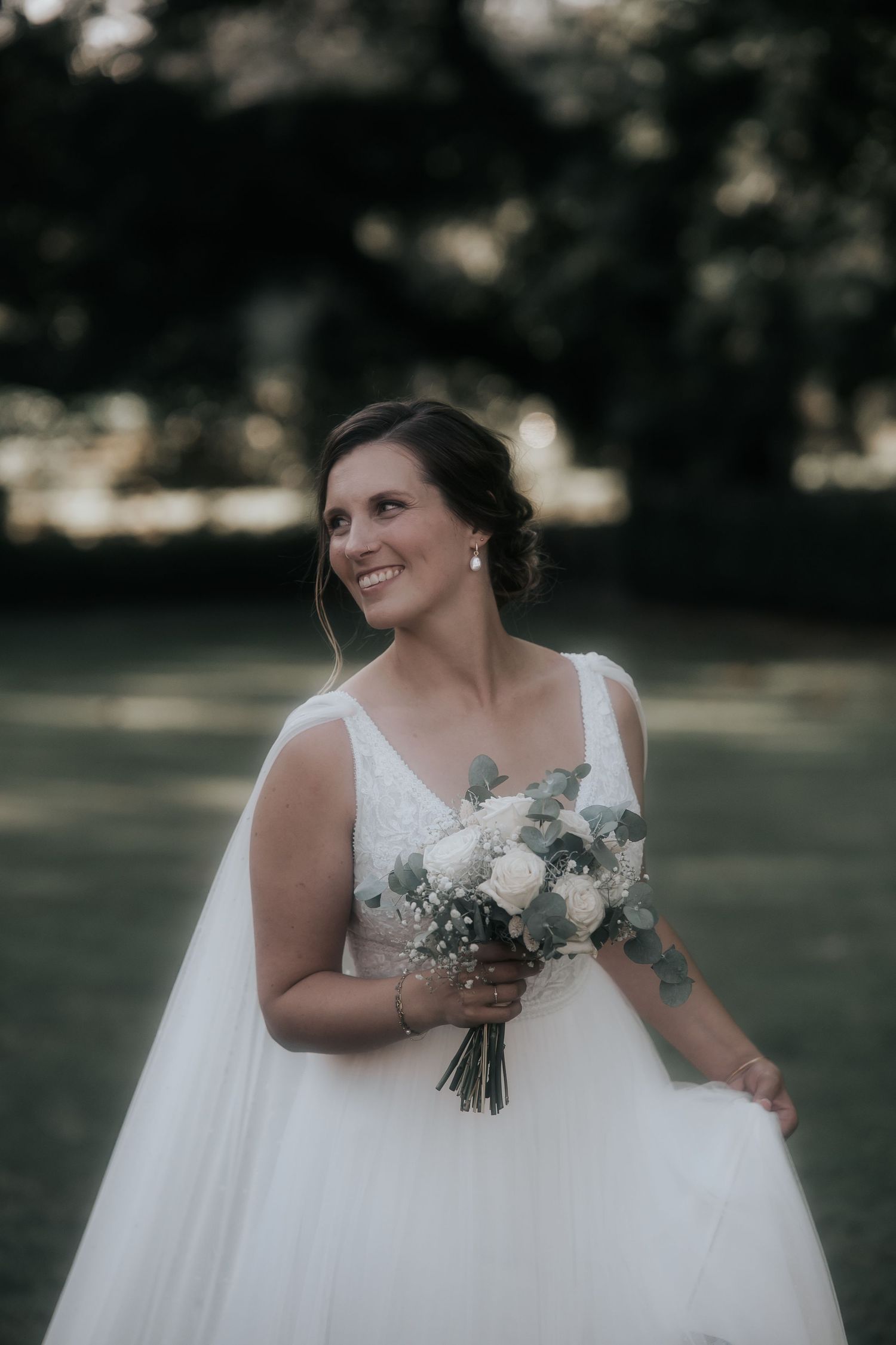 A bride in a white wedding dress holds a bouquet of white flowers while standing outdoors in a garden setting.