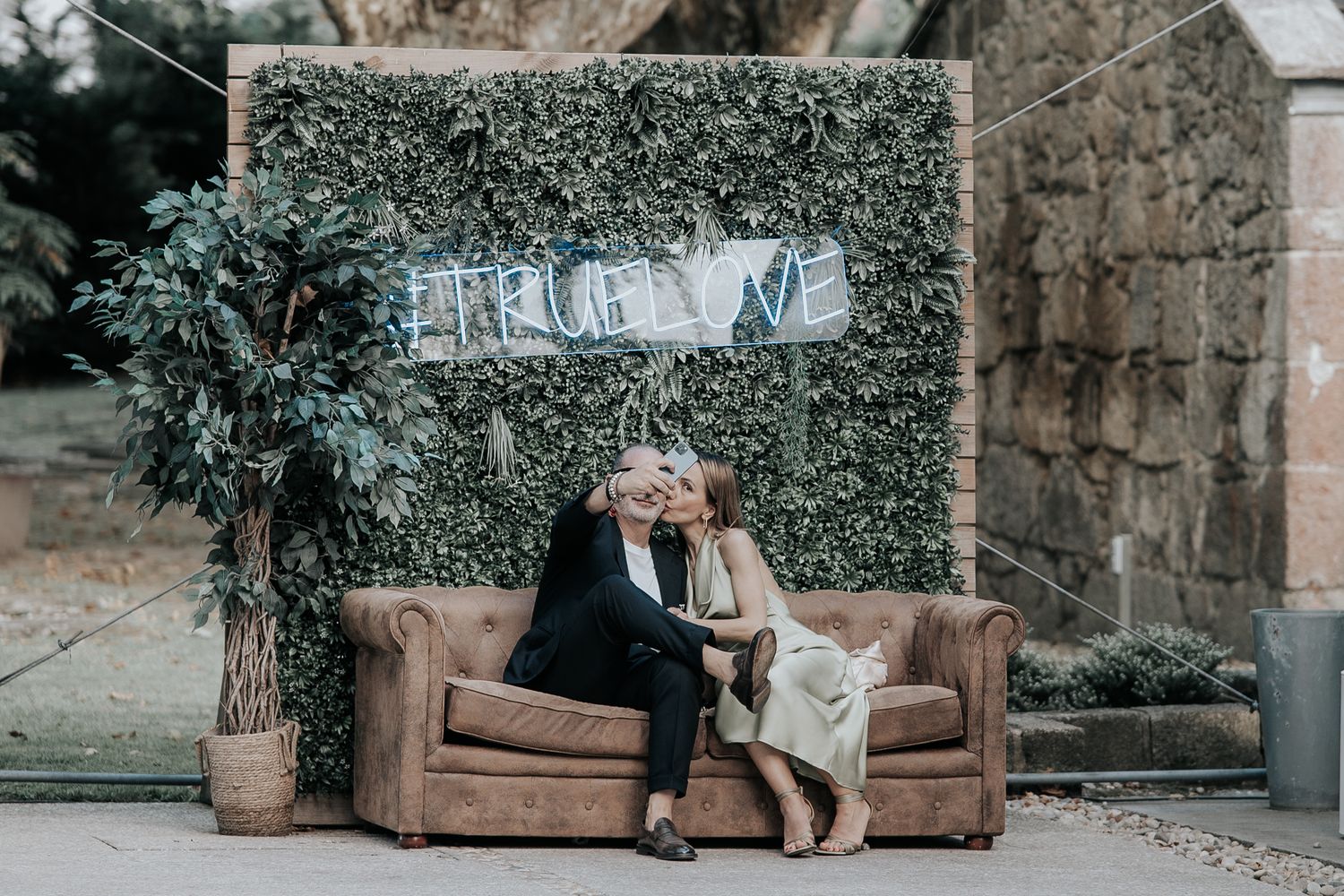 A romantic couple sits together on a brown leather couch in a garden setting surrounded by lush greenery.
