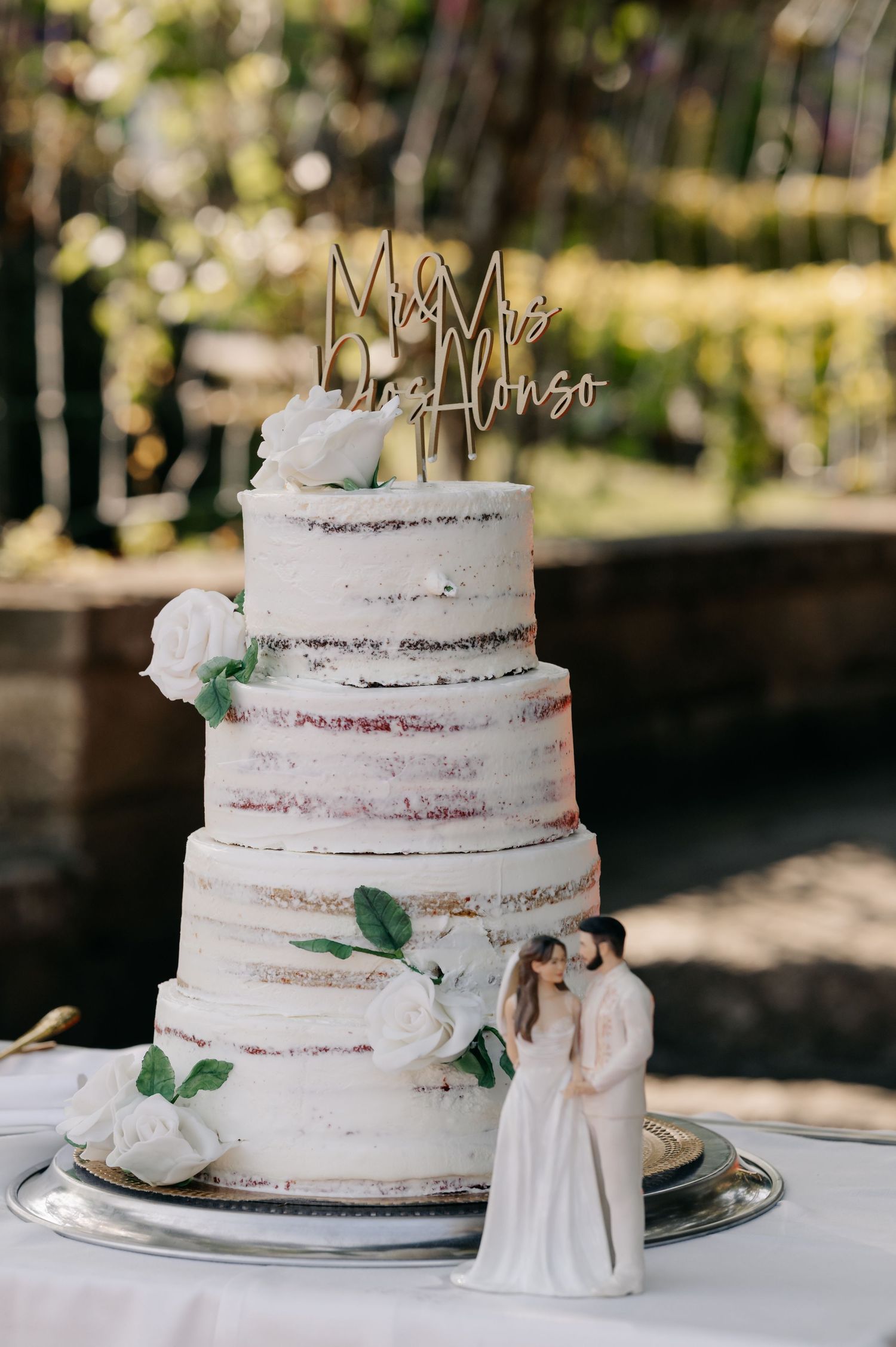 A semi-naked white wedding cake with fresh flowers and a cake topper displayed in a series of detail shots outdoors.