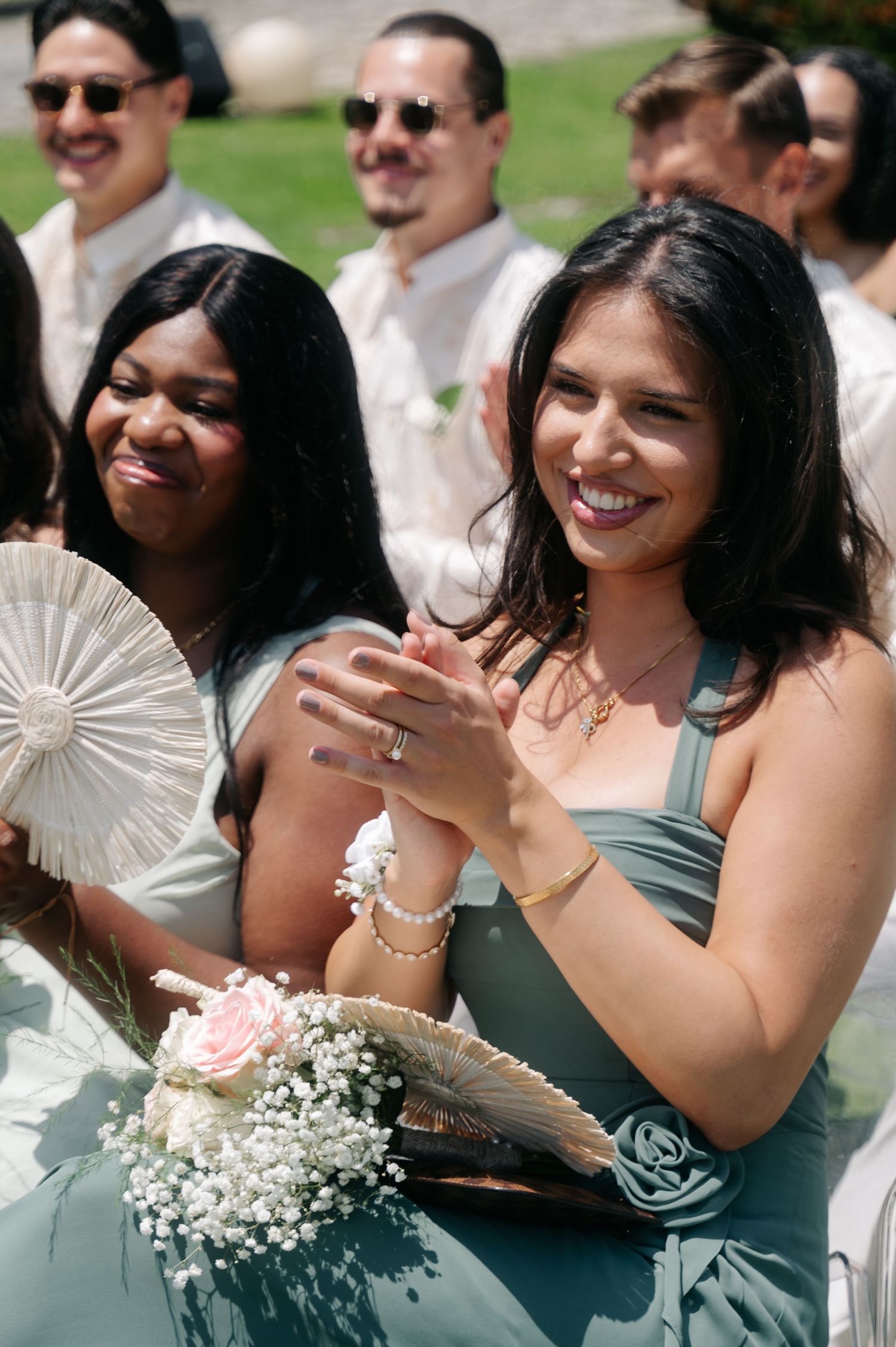 Bridesmaids in sage green dresses smile while holding fans and flowers at an outdoor wedding celebration.