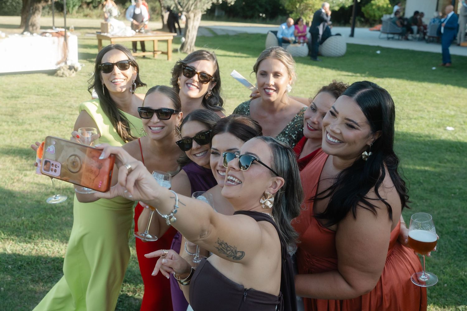 A group of friends taking a selfie together at an outdoor event while holding drinks and smiling on a sunny day.