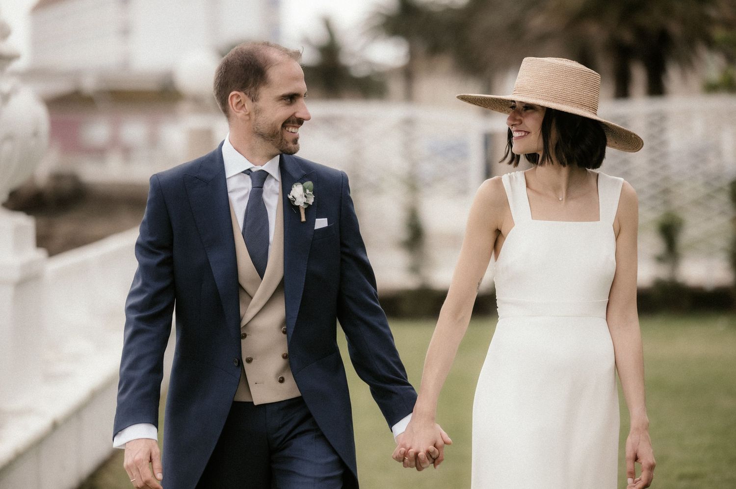 A couple in wedding attire walks hand in hand outdoors, with the bride wearing a wide-brimmed hat and simple white dress.
