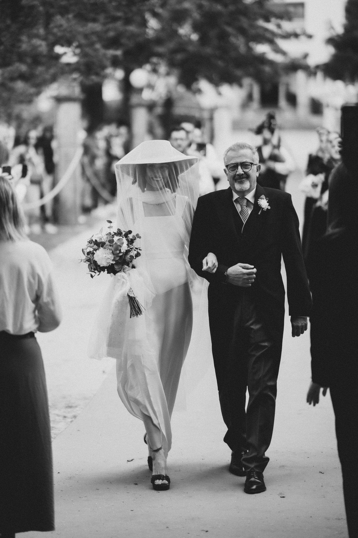 A black and white wedding photo showing a bride in a wide-brimmed hat walking arm in arm with an older gentleman.