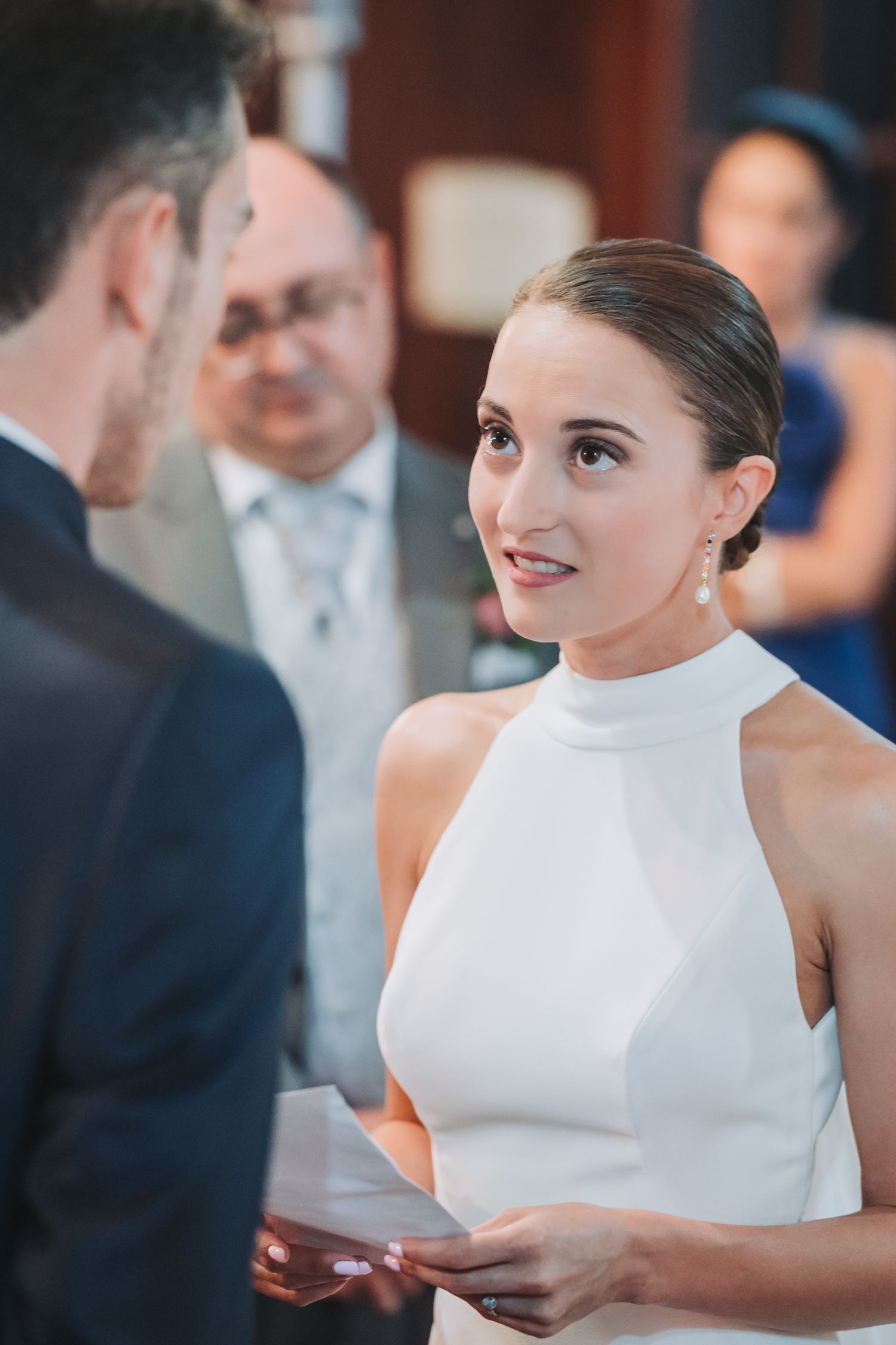 Bride in elegant white high-neck sleeveless dress reads a note during an emotional wedding ceremony moment.