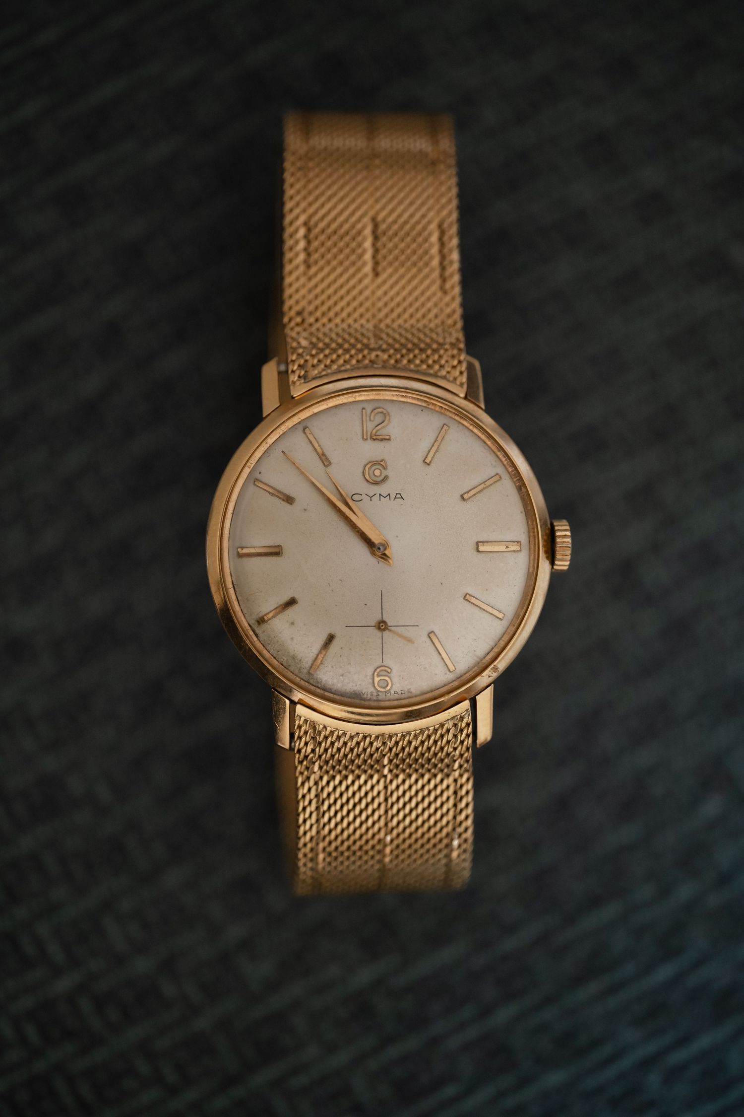 Vintage gold Cyma wristwatch with mesh band and white dial displayed on dark textured background.