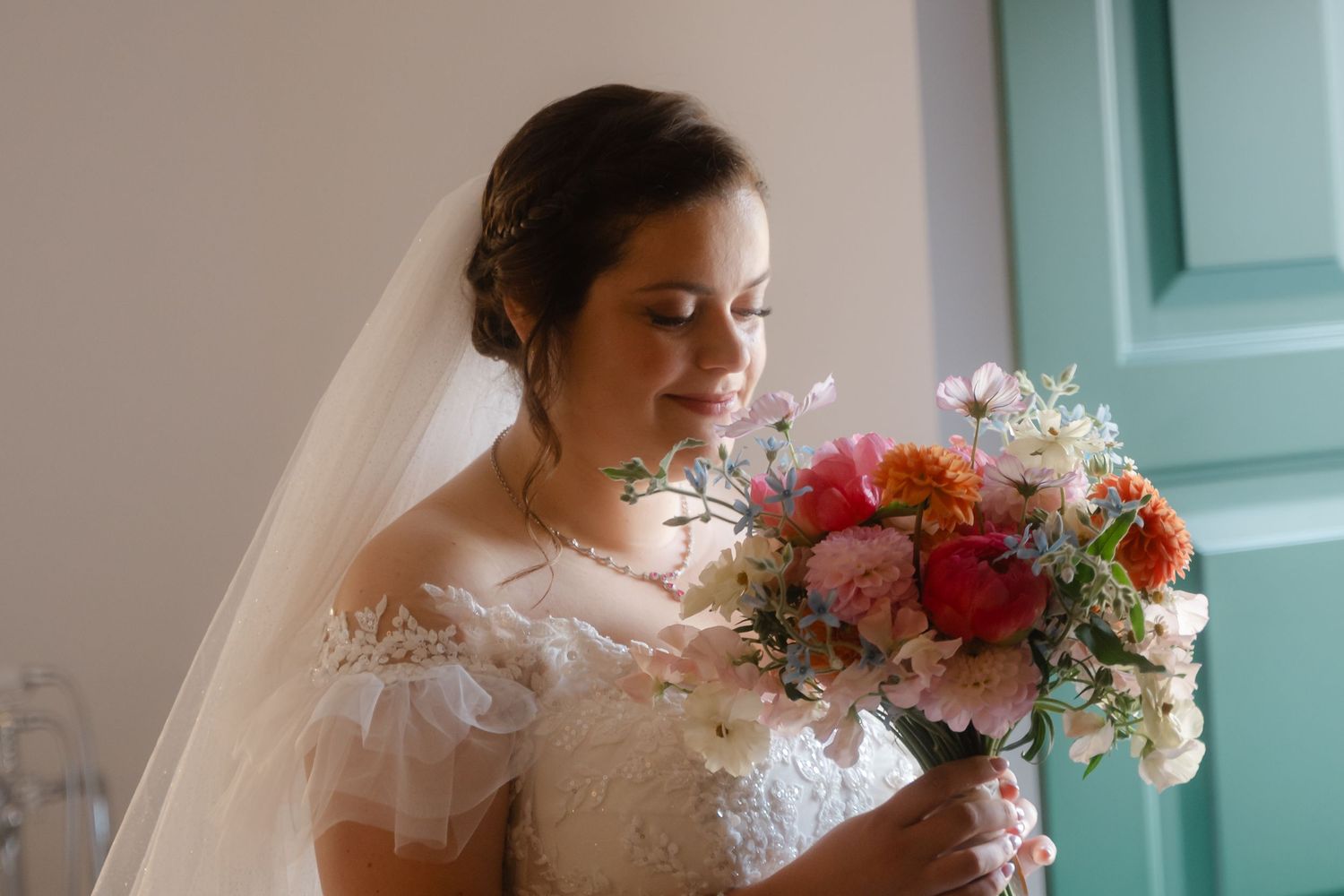 Bride in a lace wedding gown holds a vibrant bouquet of pink and red flowers against a turquoise wall.
