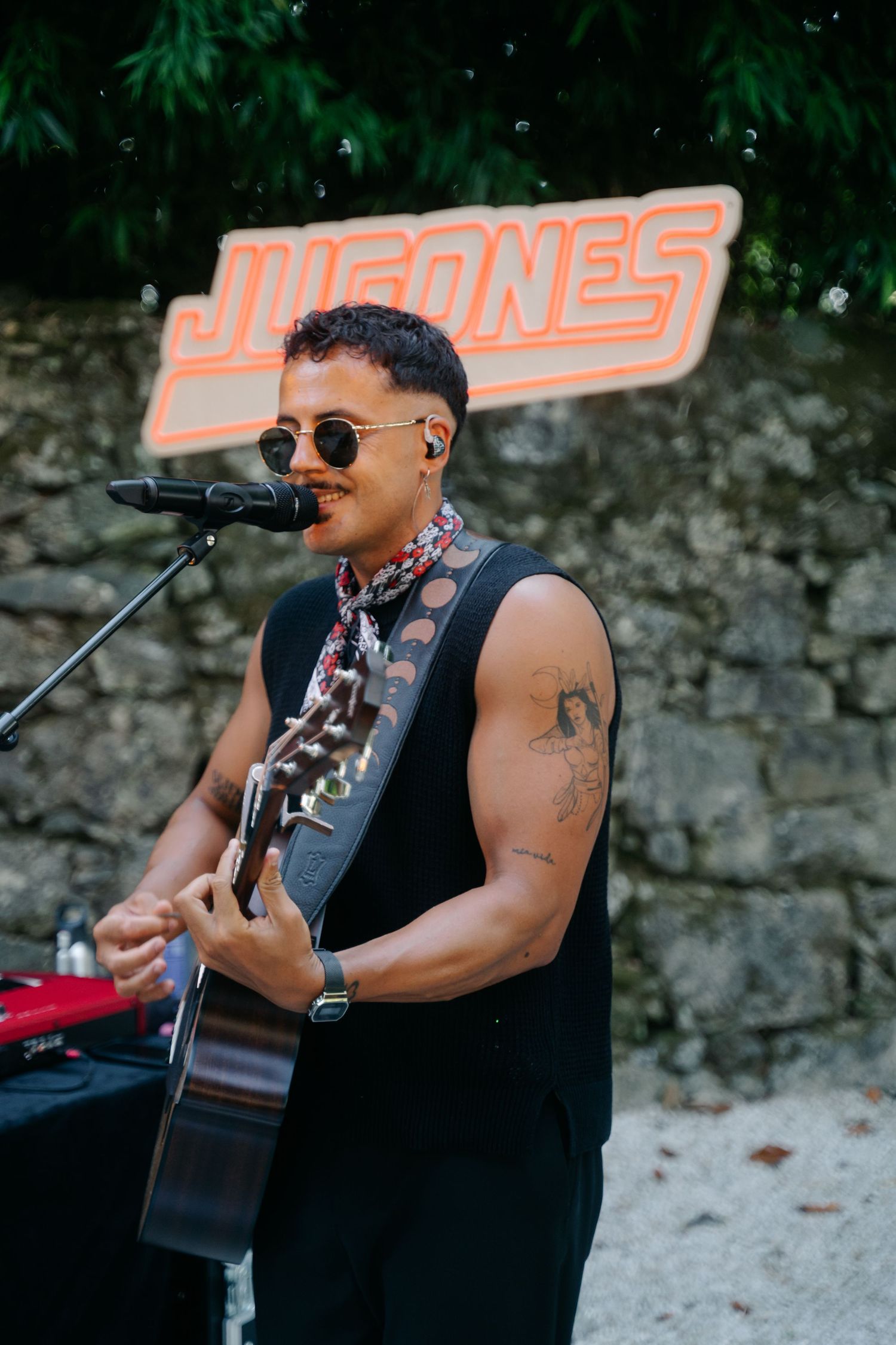 A musician in a black tank top performs with an acoustic guitar outdoors in front of an orange Juanes sign.