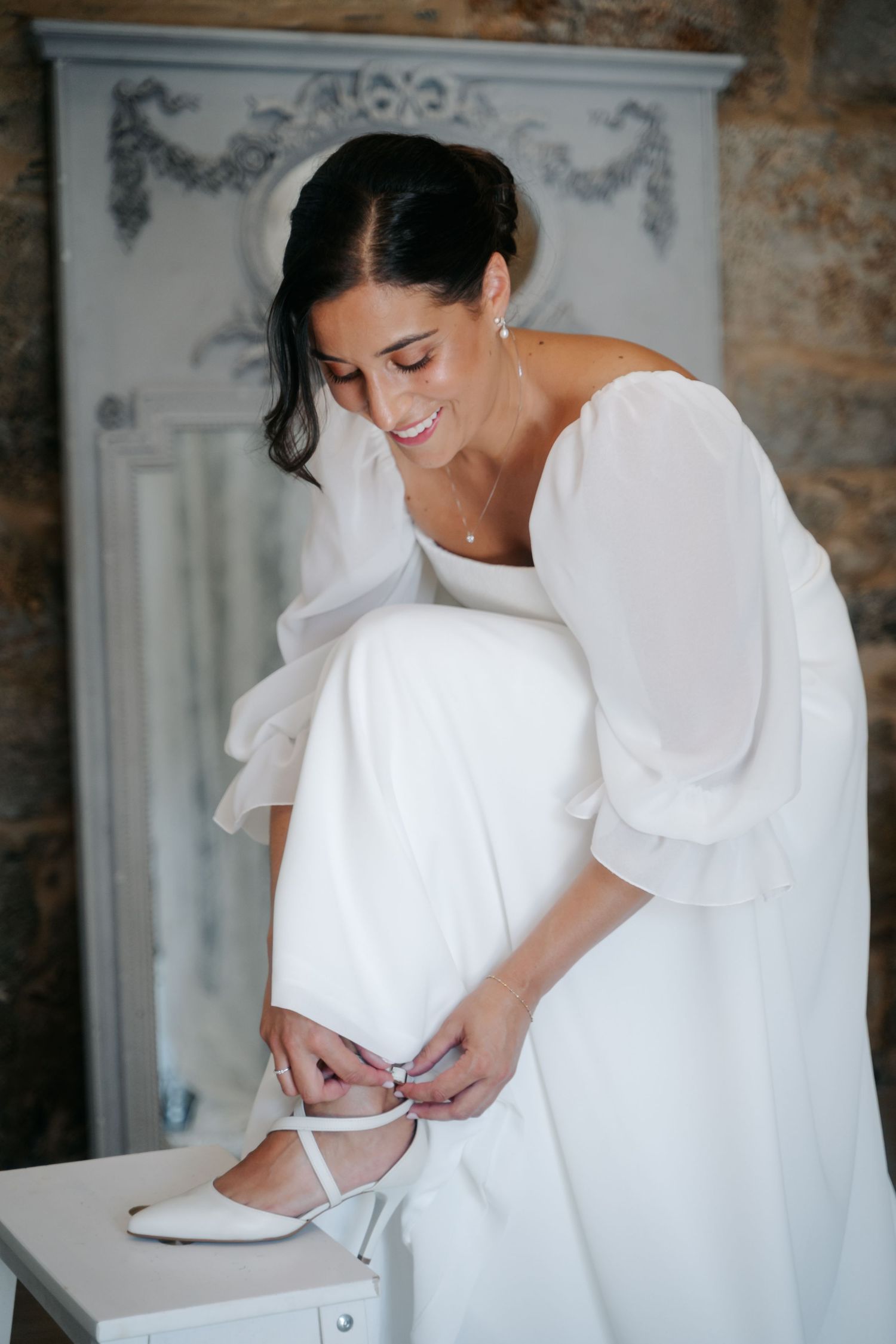 Bride in a white wedding dress adjusts her gown while standing in an elegant room with exposed brick walls.