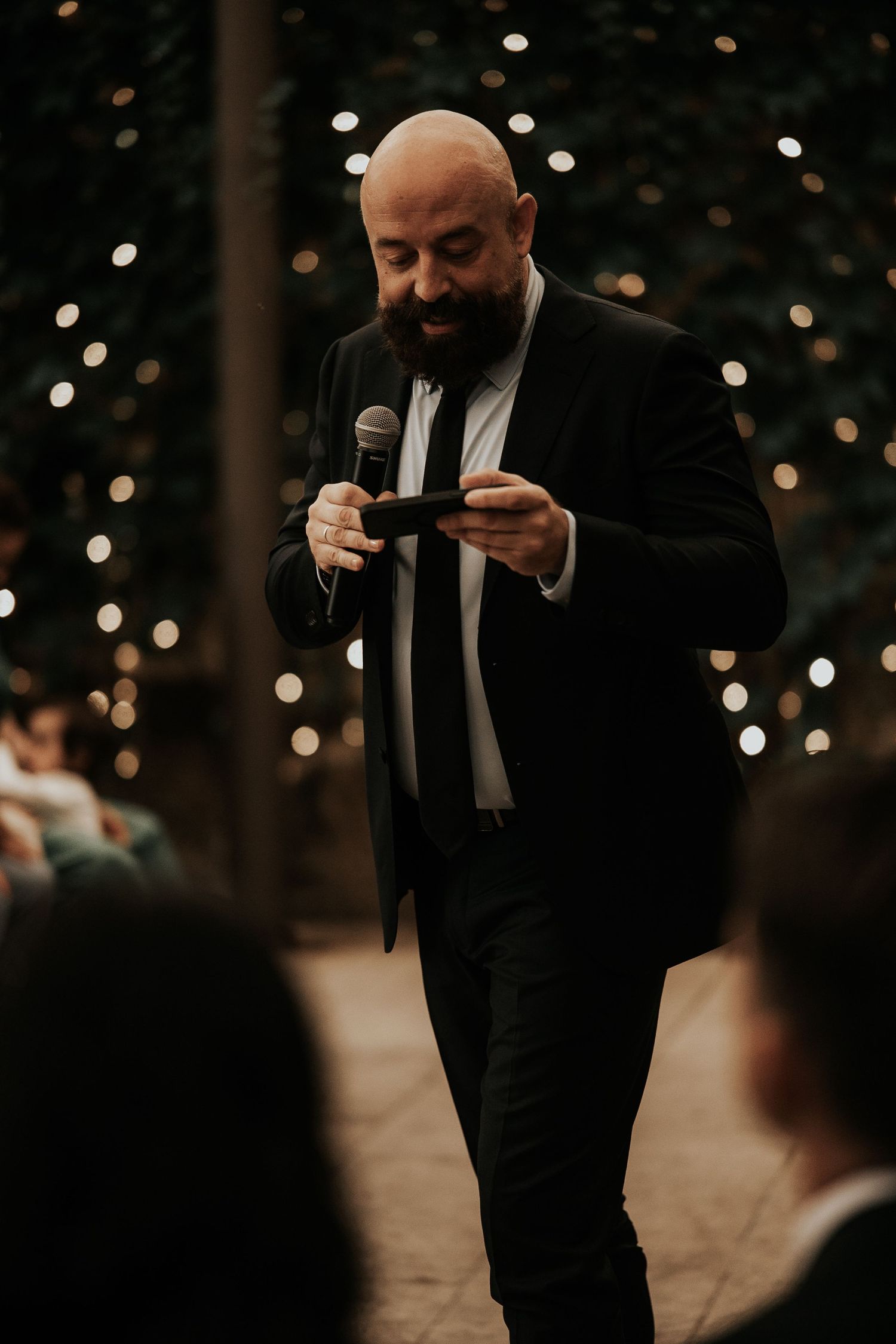 Person in black suit speaking into microphone against bokeh lights background.