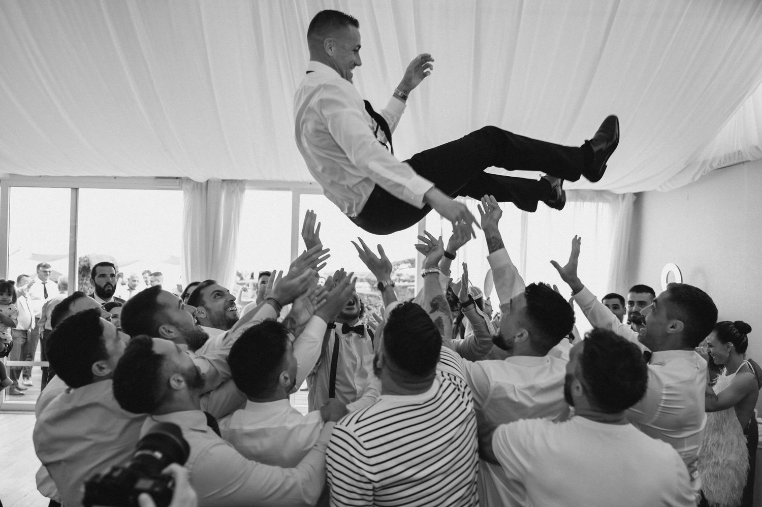 Black and white photo of wedding guests tossing the groom in the air during a joyful celebration.