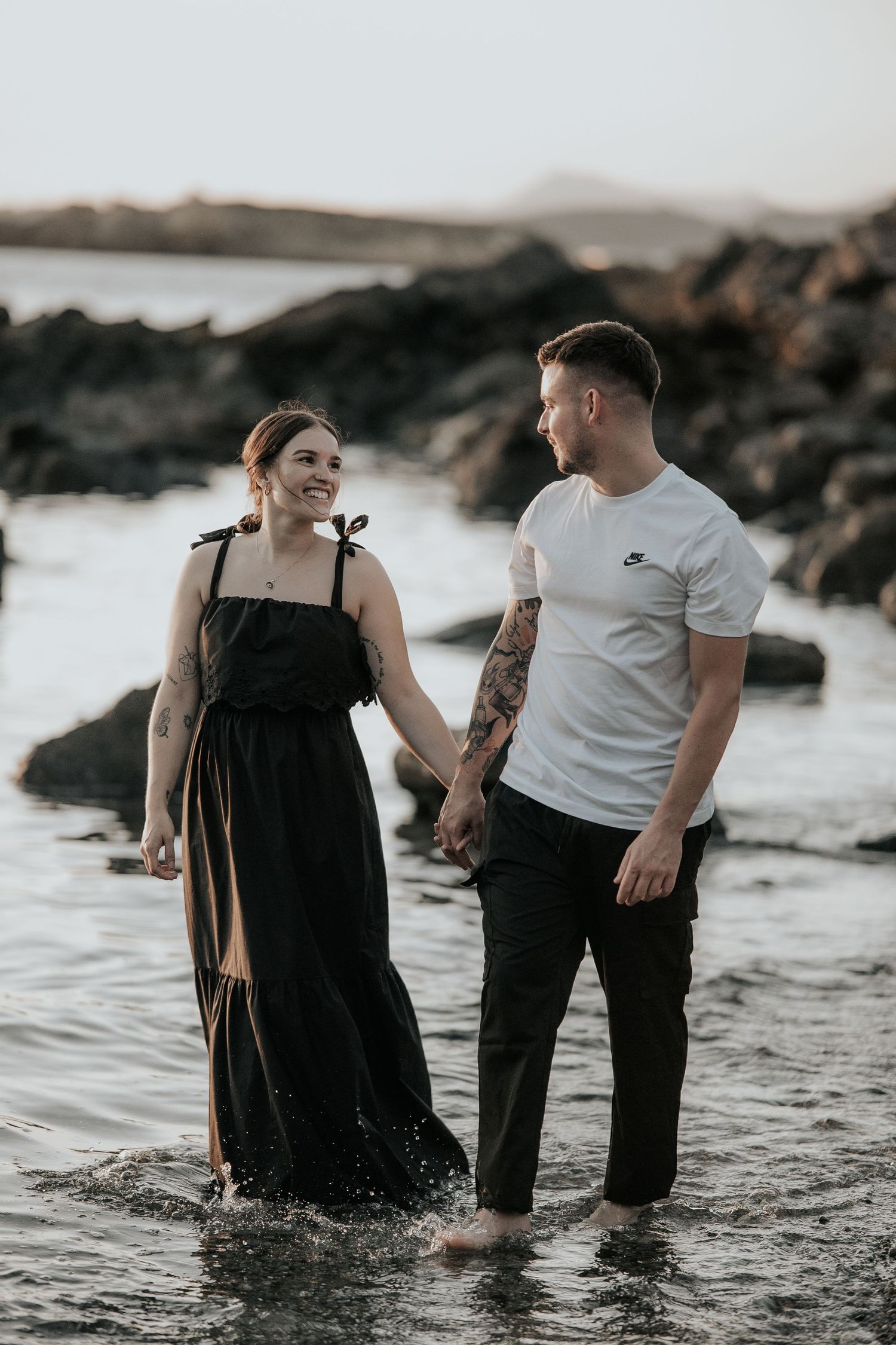A couple stands in shallow ocean water during sunset while wearing black and white outfits at the beach.
