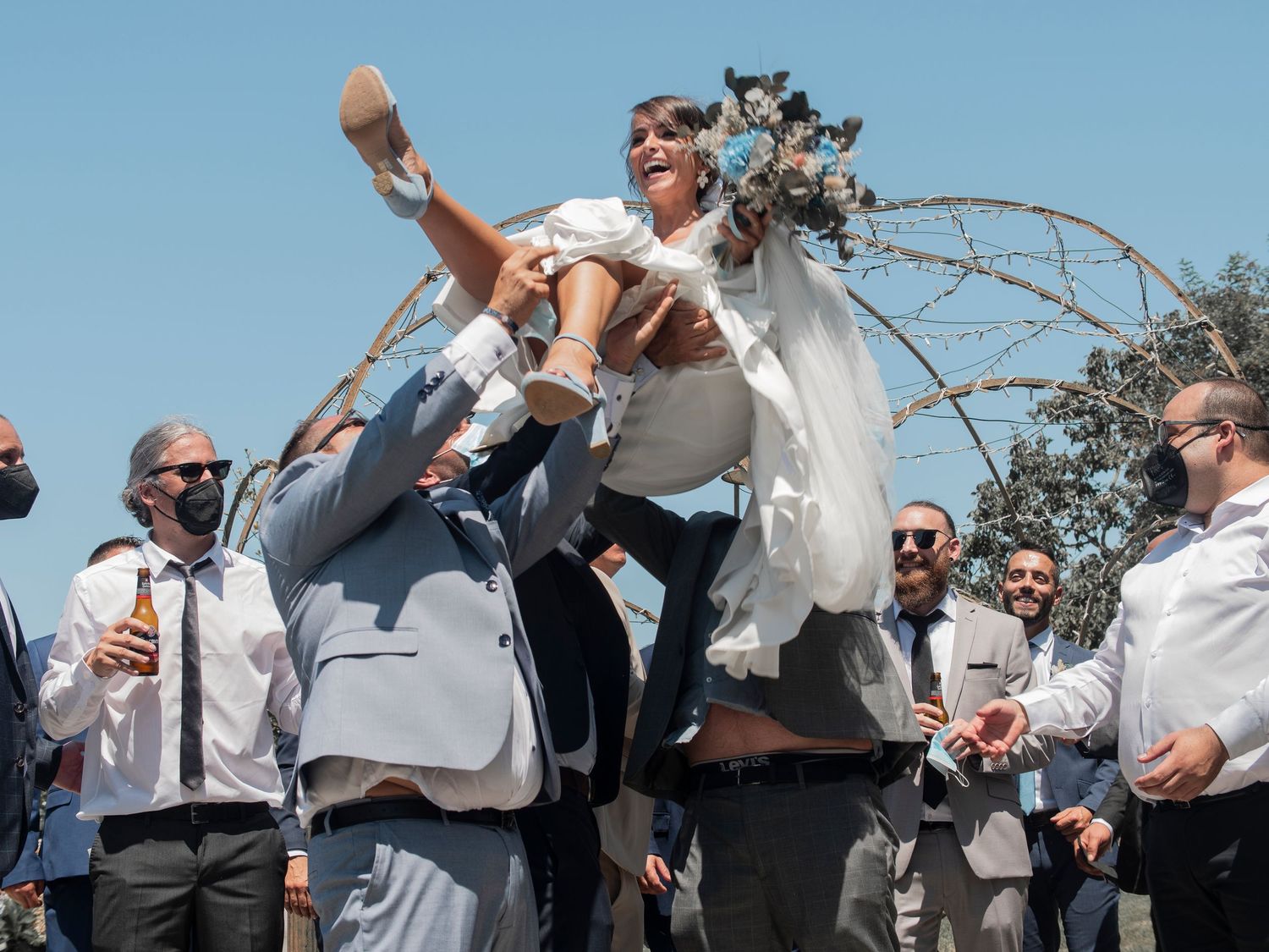 A group of wedding guests in white shirts celebrate outdoors under a bright blue sky.