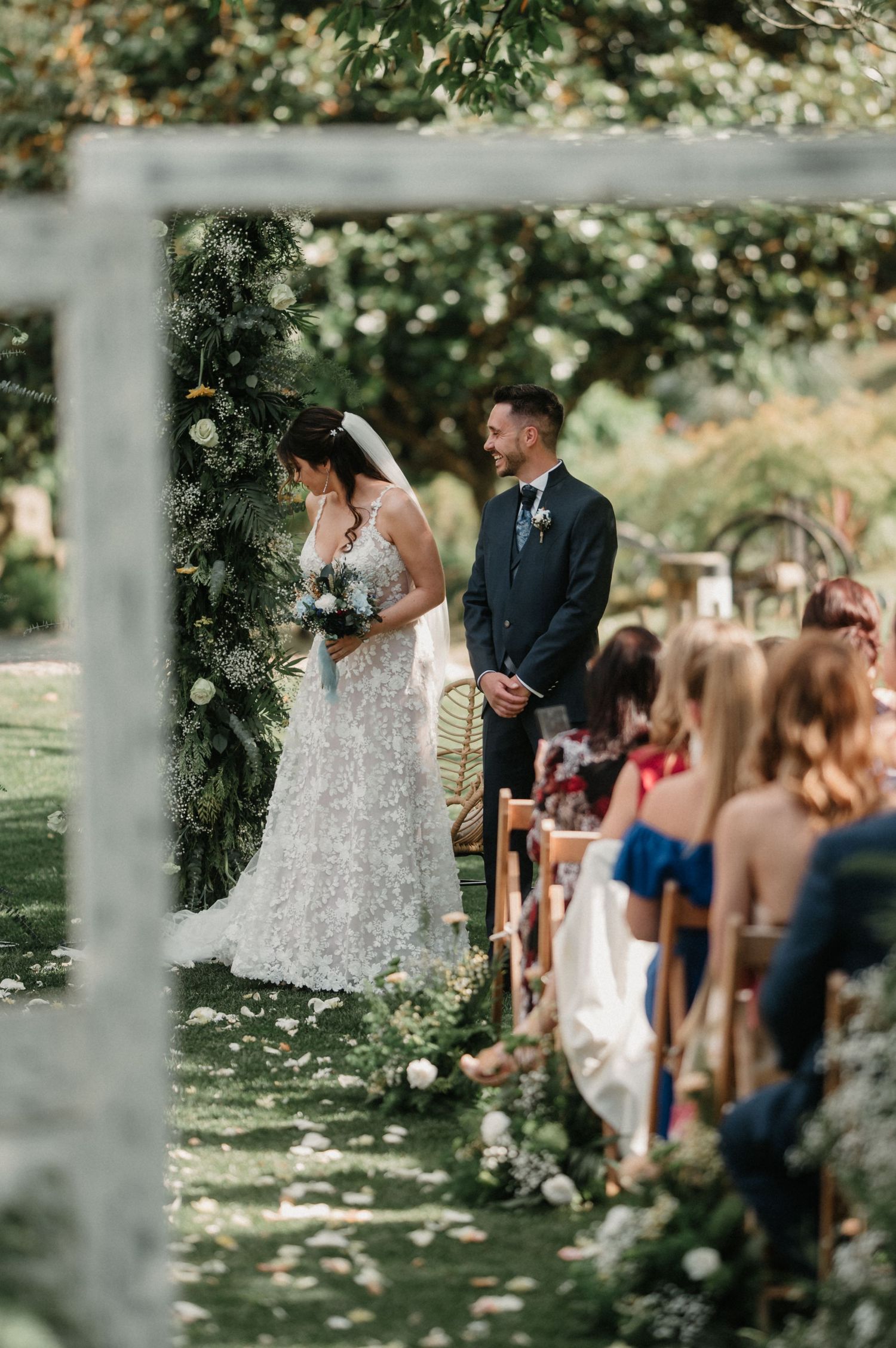 Outdoor wedding ceremony under a floral arch with guests seated on wooden chairs.