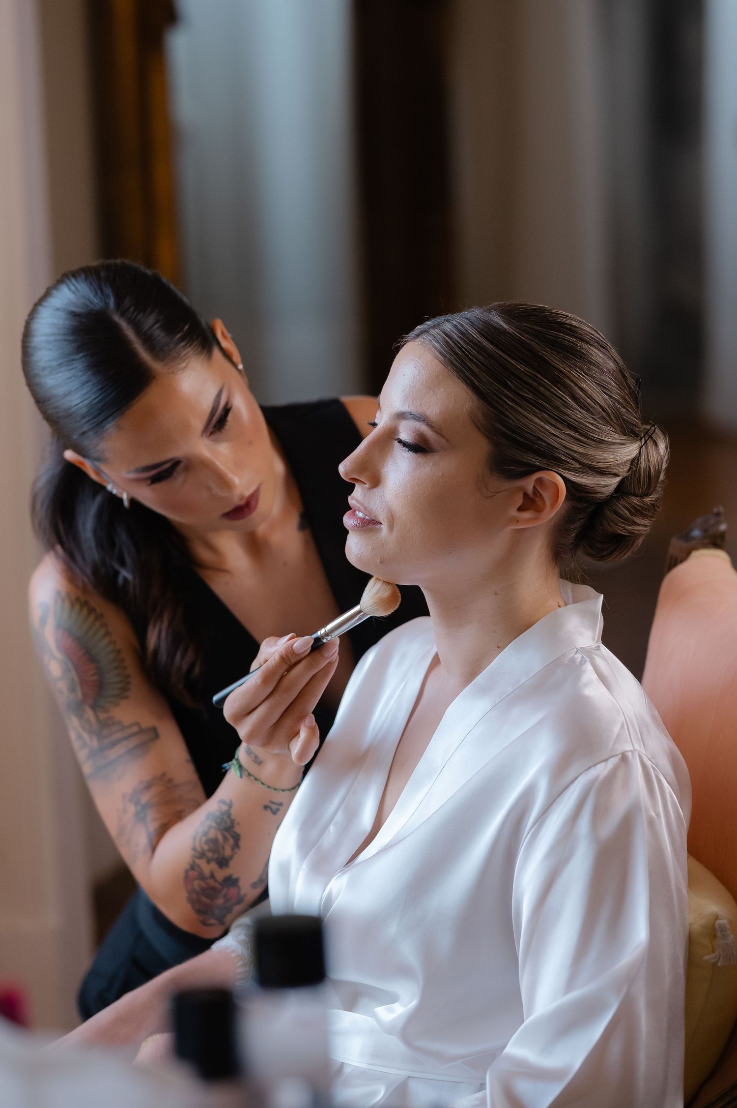 Makeup artist with tattooed arms applies cosmetics to client wearing white silk robe during beauty preparation session.