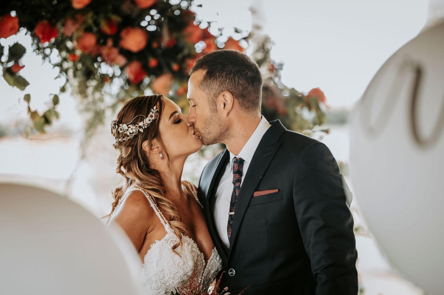 Romantic wedding couple shares intimate kiss under floral arch decorated with coral and peach blooms.
