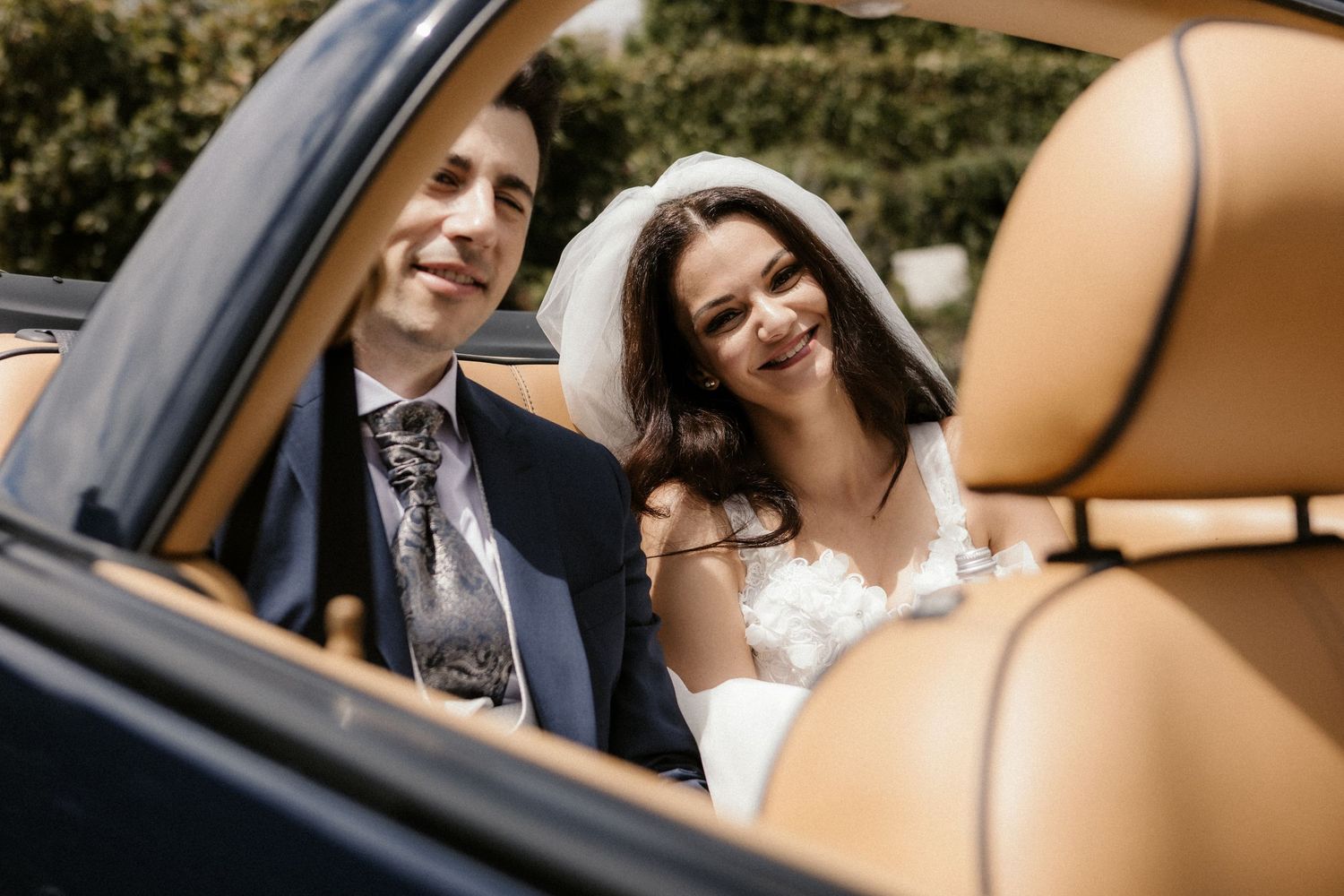 Happy newlyweds smile together while sitting in a luxury car with tan leather seats.