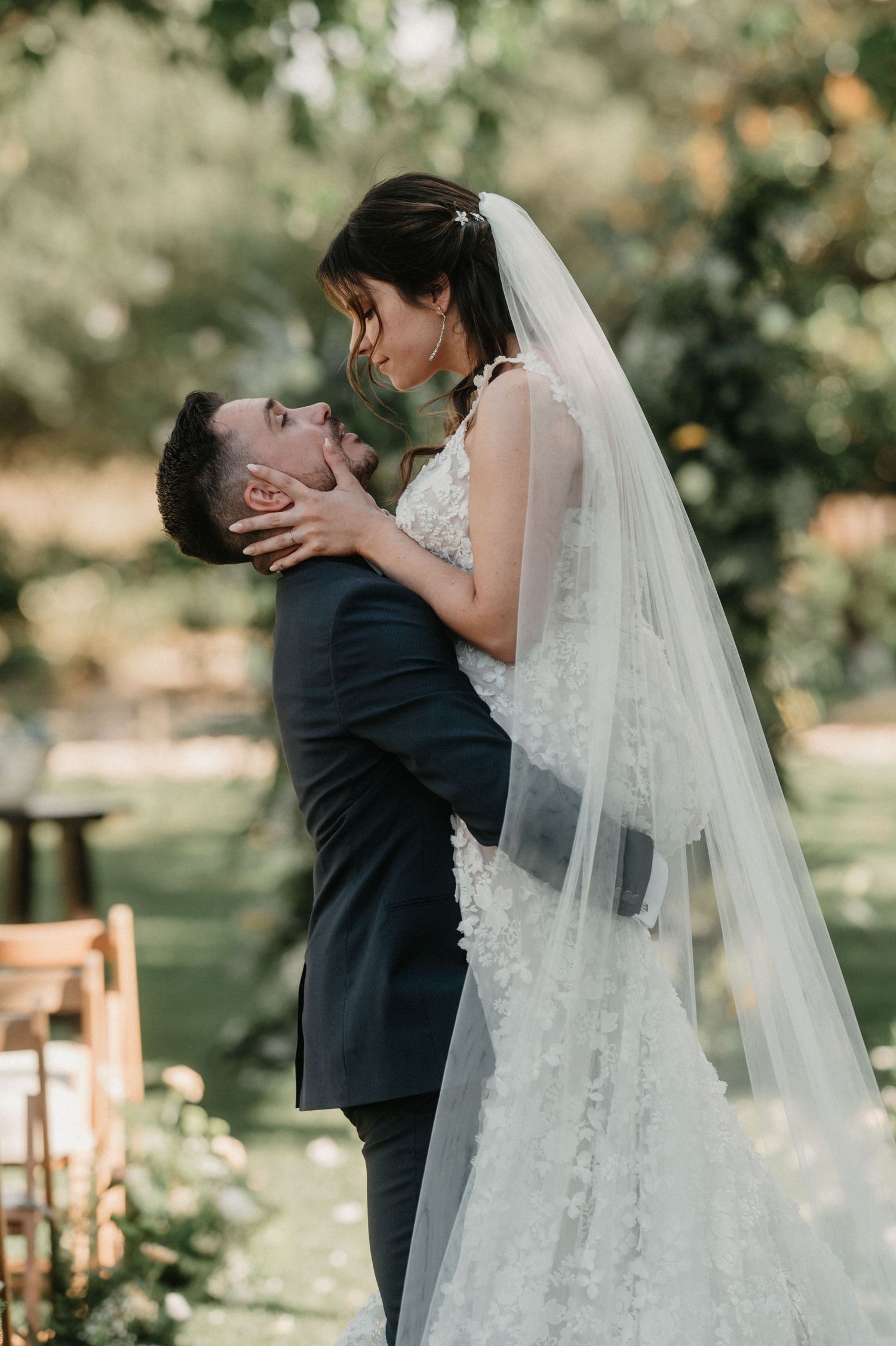 A romantic wedding couple shares tender kisses in a sunlit garden setting while the bride's lace veil flows beautifully.