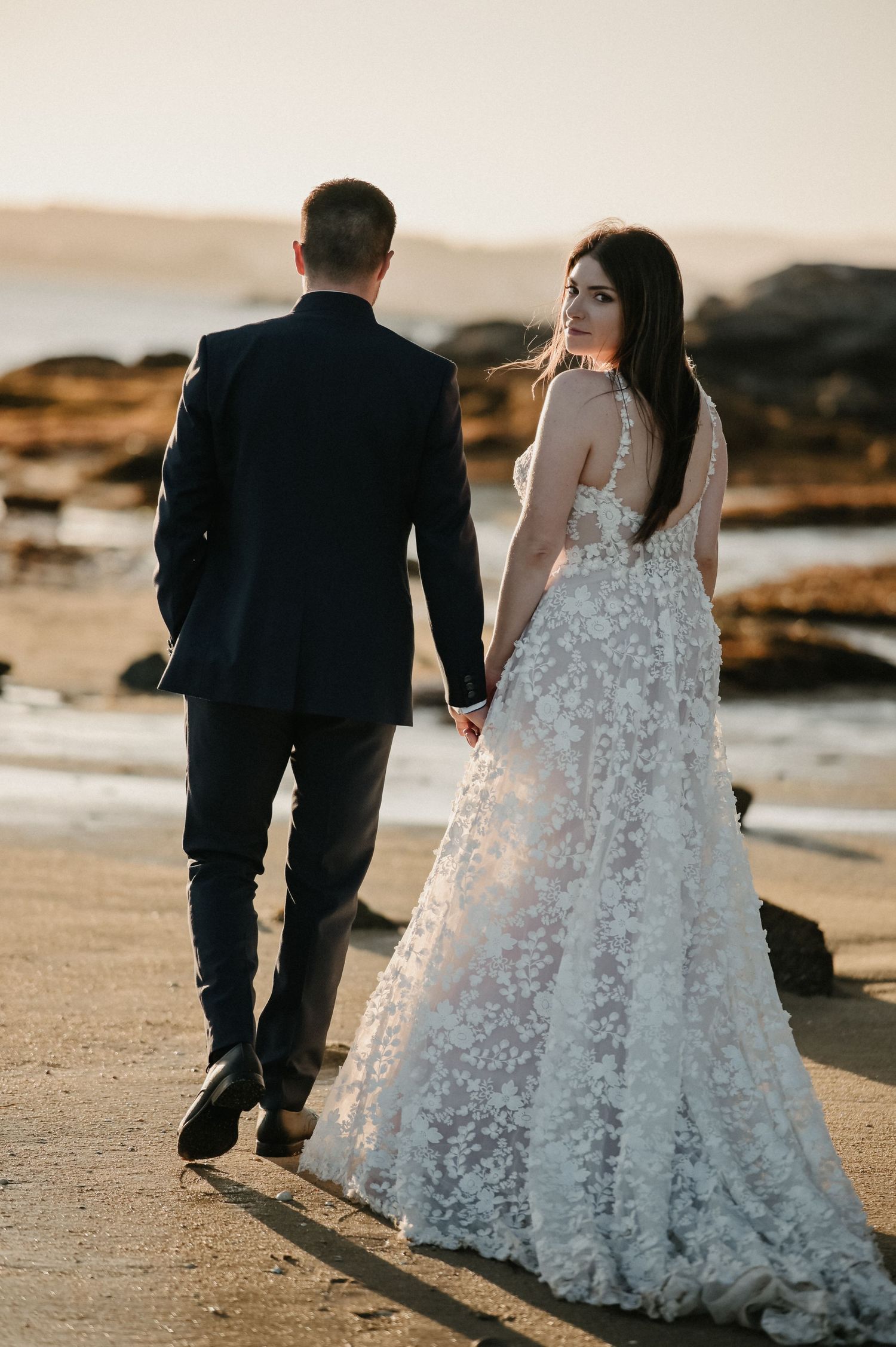 Newlyweds walking together on a beach at sunset, bride wearing a detailed lace wedding gown.