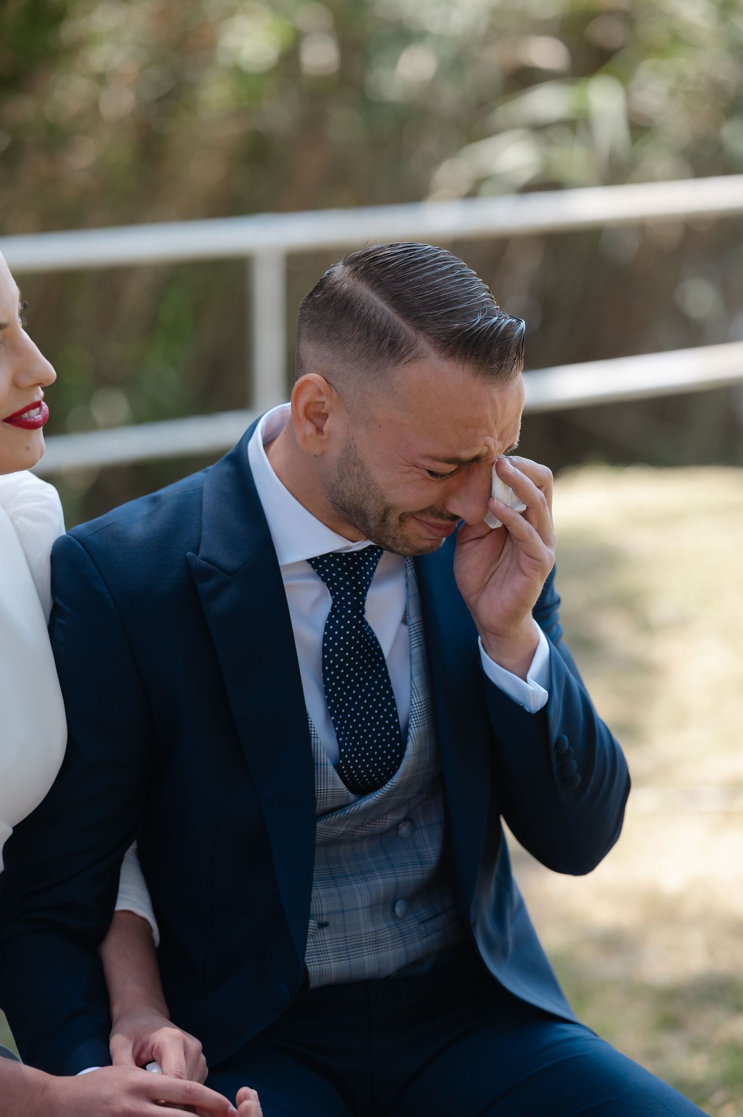 Emotional groom in navy suit wipes tears during outdoor wedding ceremony.