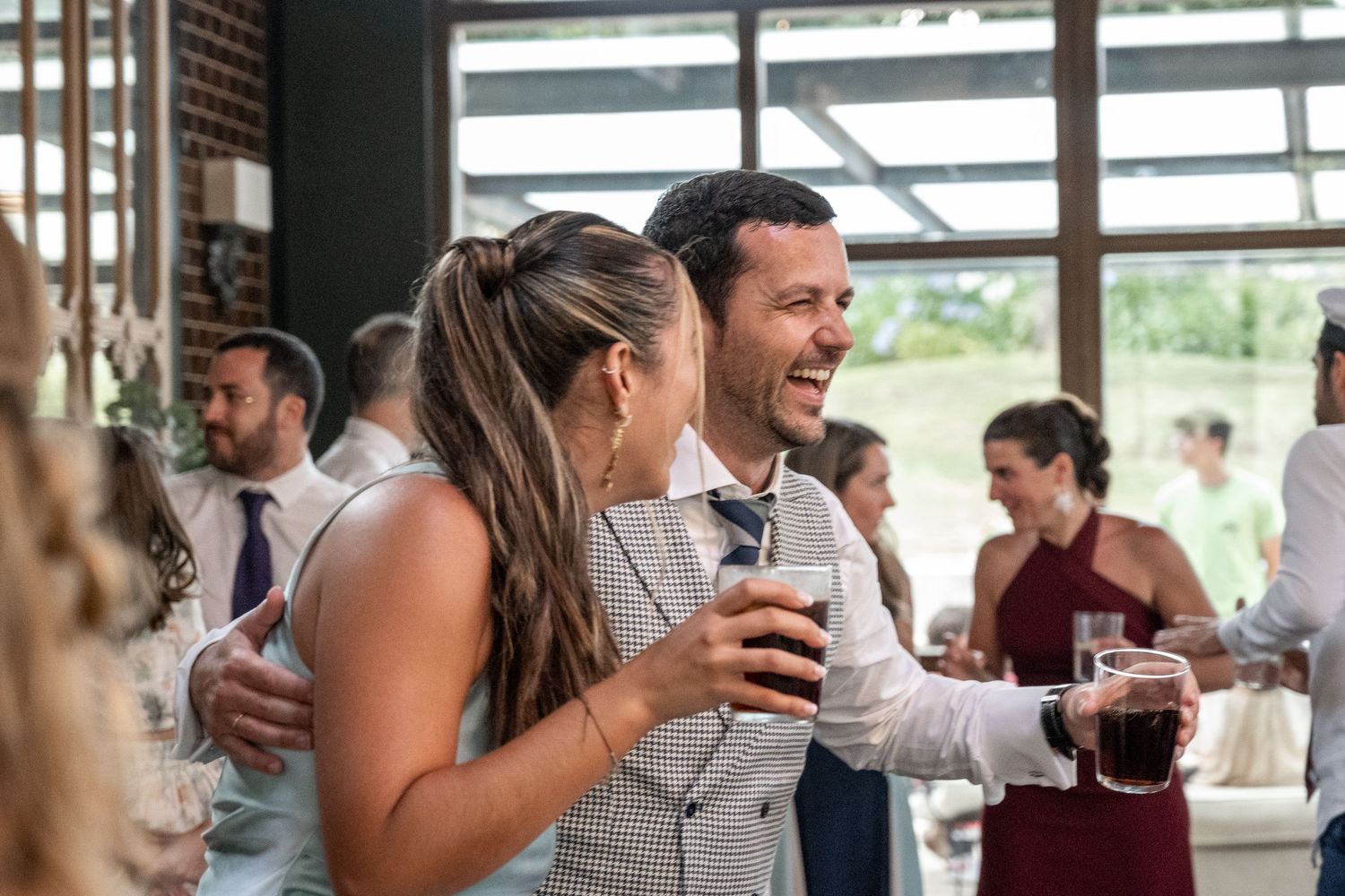 Wedding guests laugh and socialize while holding drinks at an indoor reception venue.