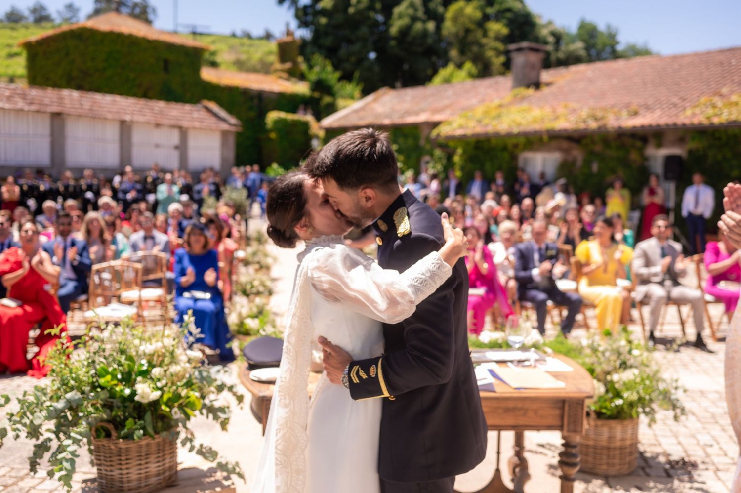 Wedding guests gather in a sunny outdoor courtyard with Spanish-style architecture and floral decorations.