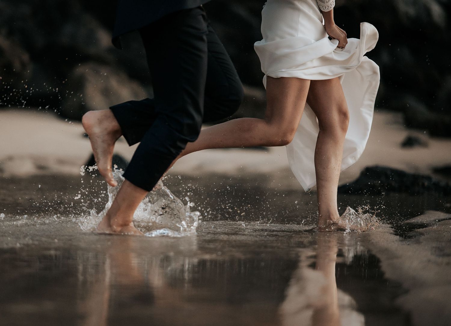 Playful couple splashing in shallow water, with white dress and formal attire creating movement.