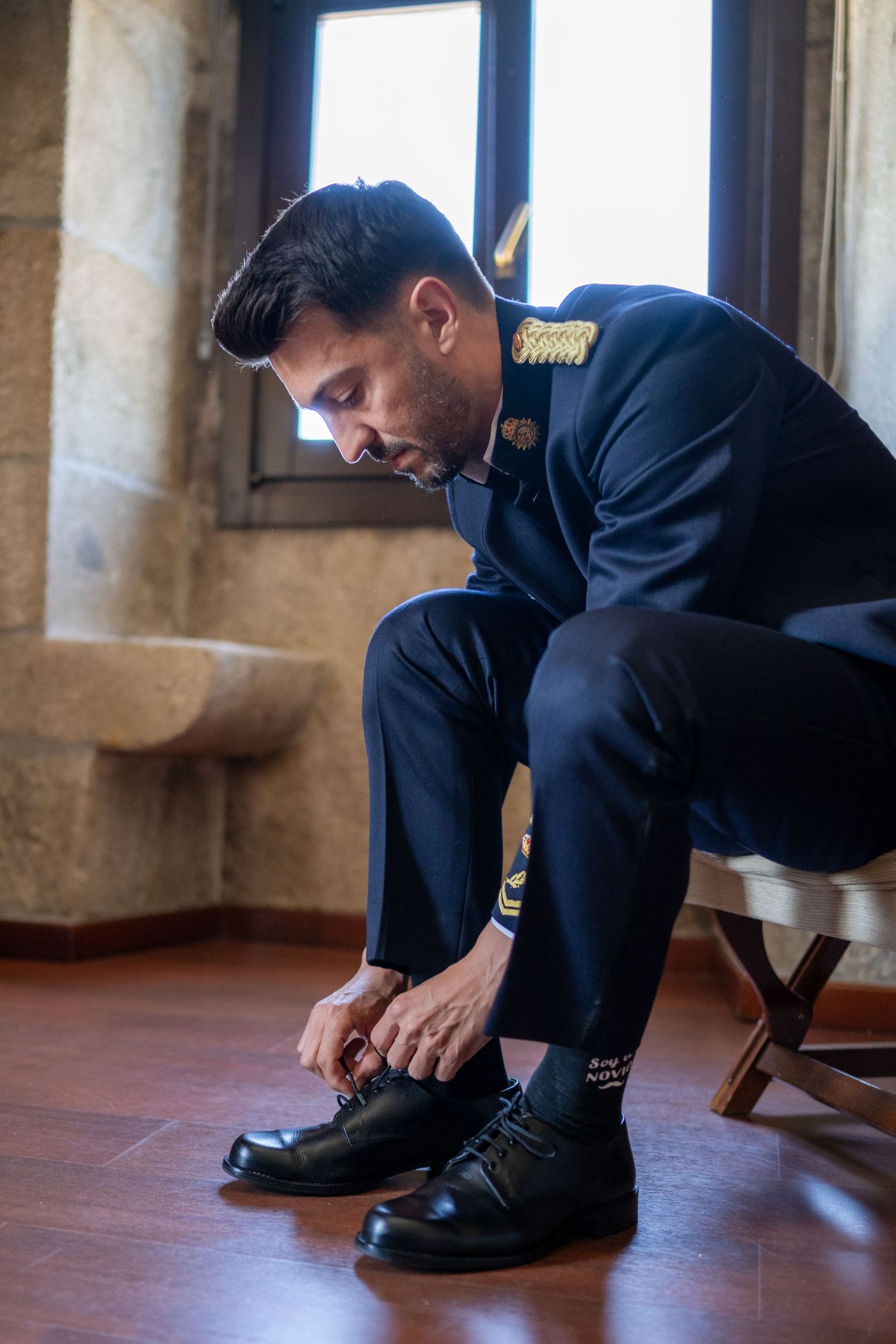 Military officer in dress uniform sits on a chair while tying formal black dress shoes on hardwood floor.
