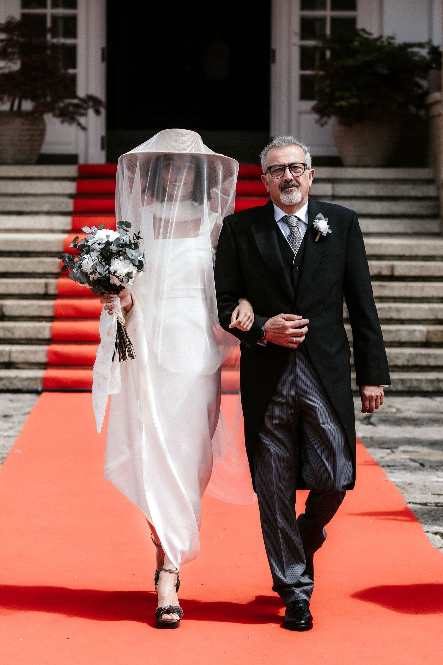 A couple walking down a red carpet with the bride in white dress and groom in black suit.