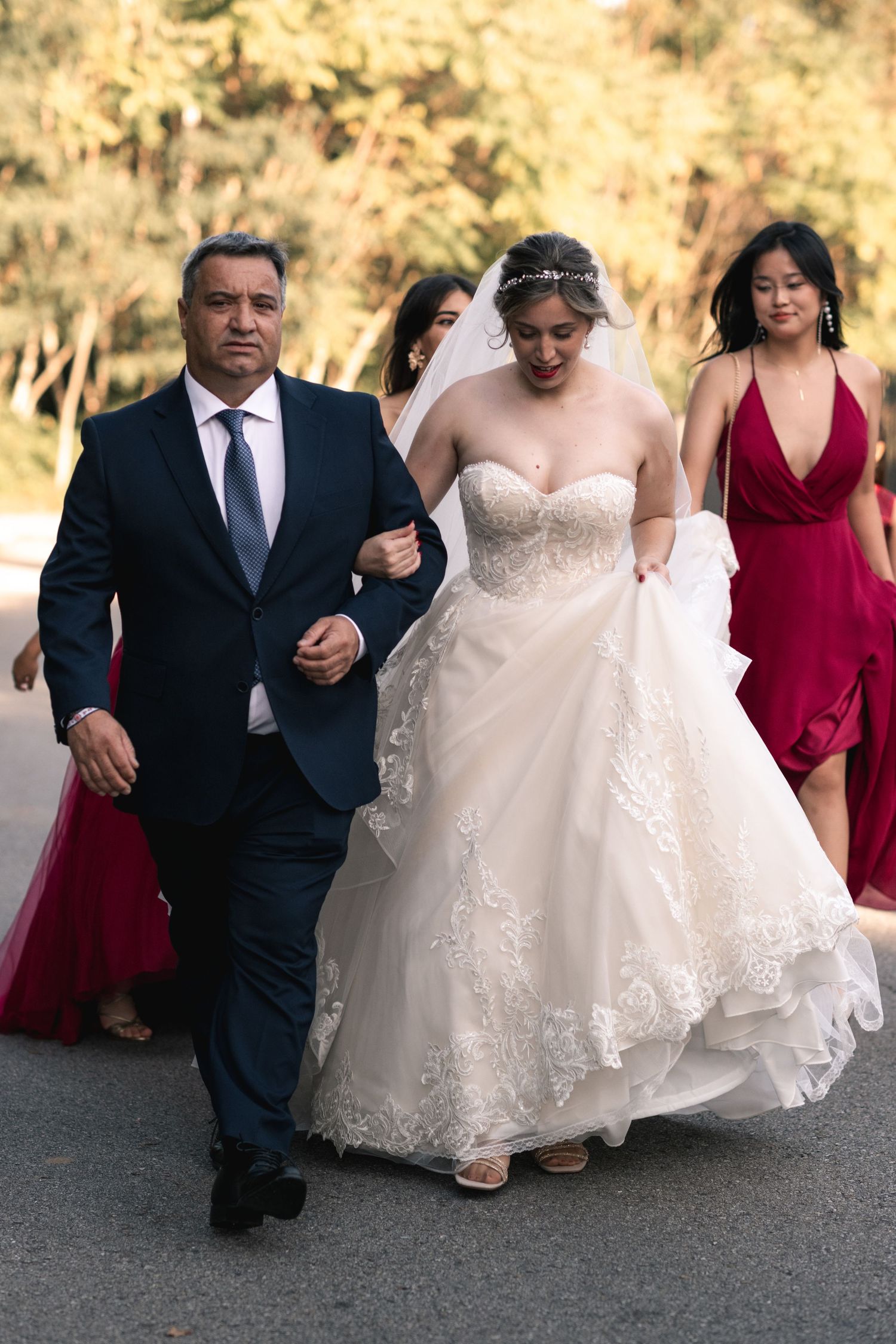 Wedding party walks together outdoors with bride in white lace gown accompanied by attendants in burgundy dresses.