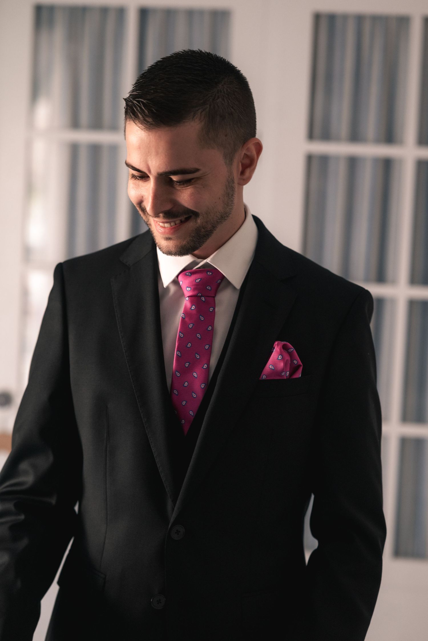 A person in a black suit with a vibrant magenta tie and pocket square smiles while looking down.