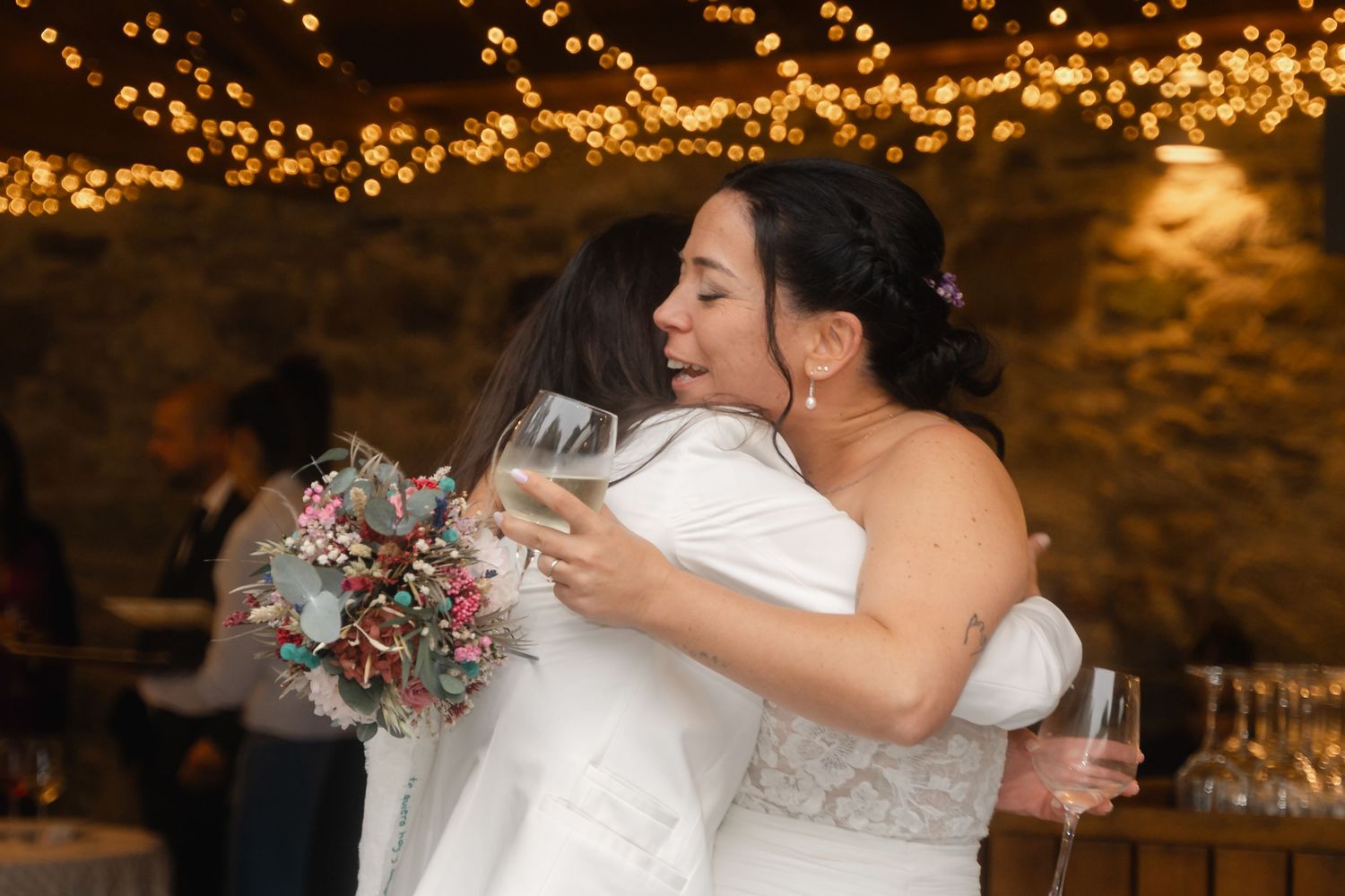 A bride in a white dress holds a bouquet and wine glass under twinkling lights at a reception.