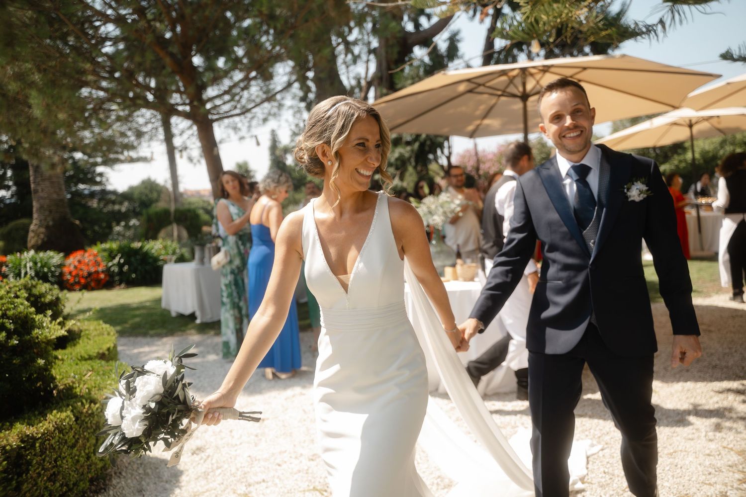 Wedding party celebrates outdoors under a beige umbrella on a sunny day.