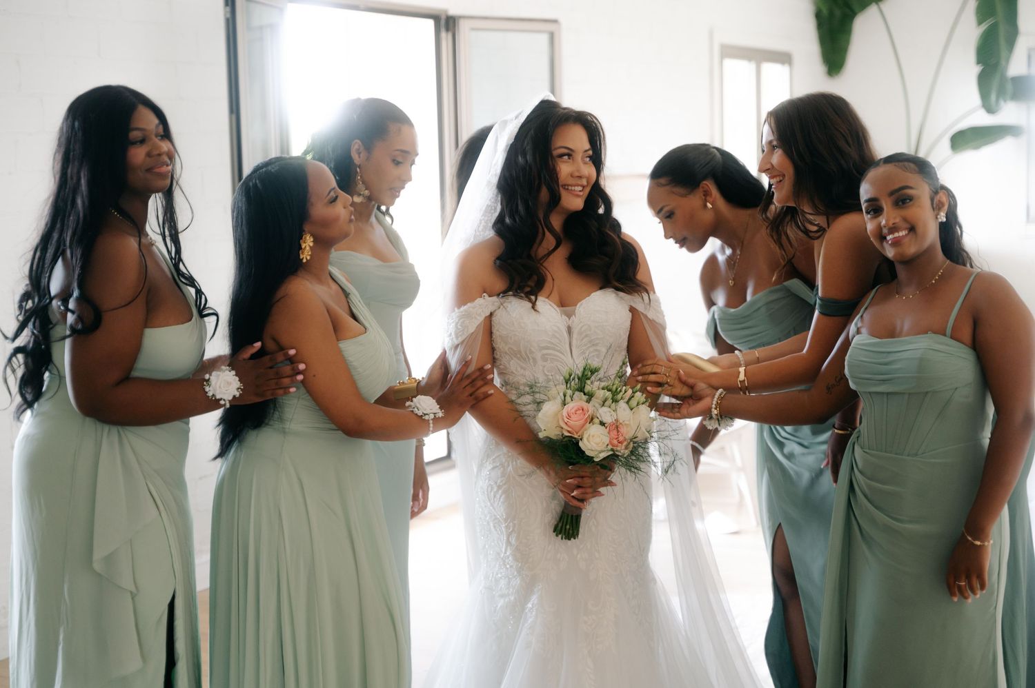 Bridesmaids in sage green dresses share joyful moments while getting ready in a sunlit room.