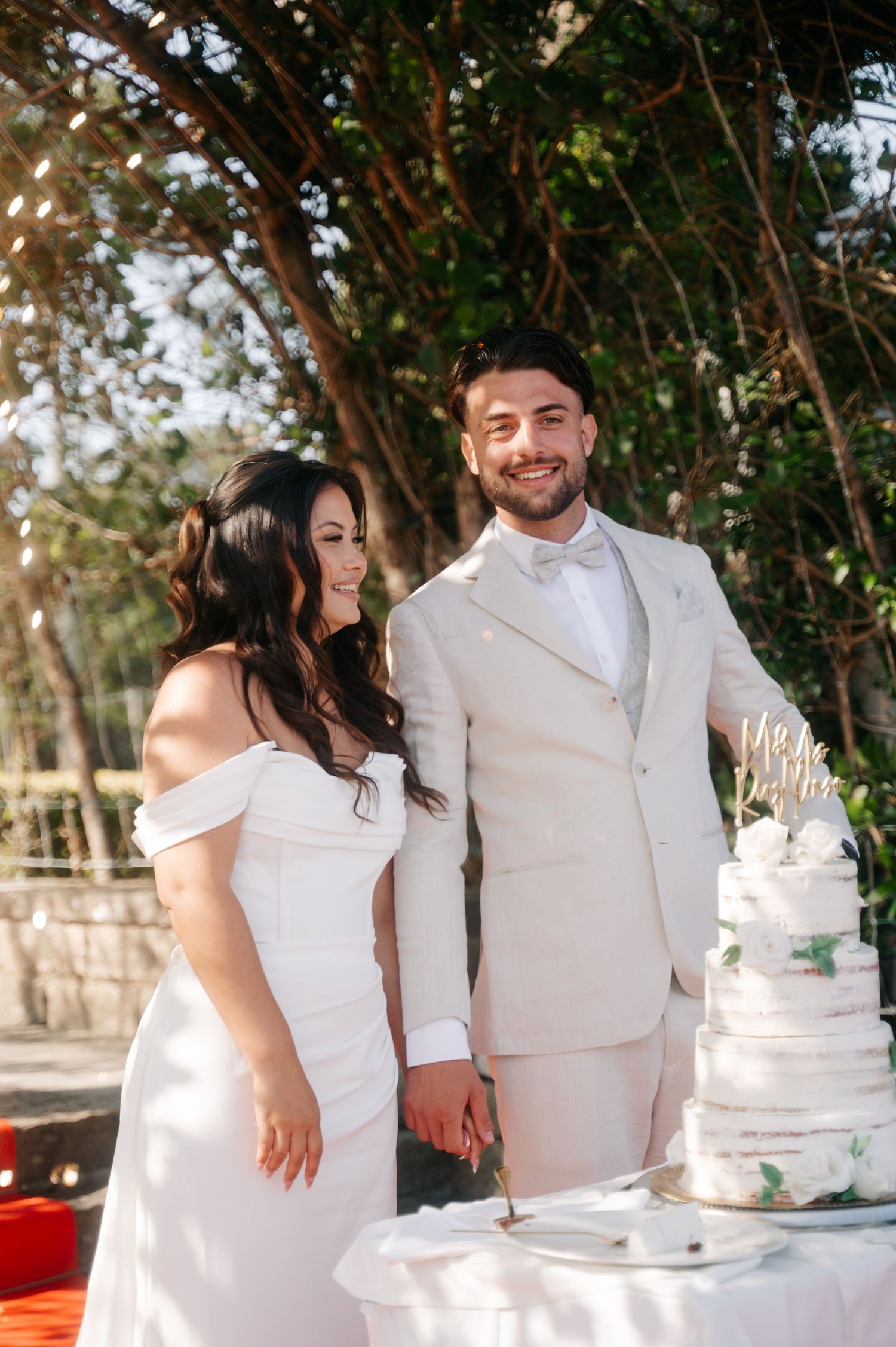 A couple in white attire stands together at their wedding reception near an elegant white tiered cake.