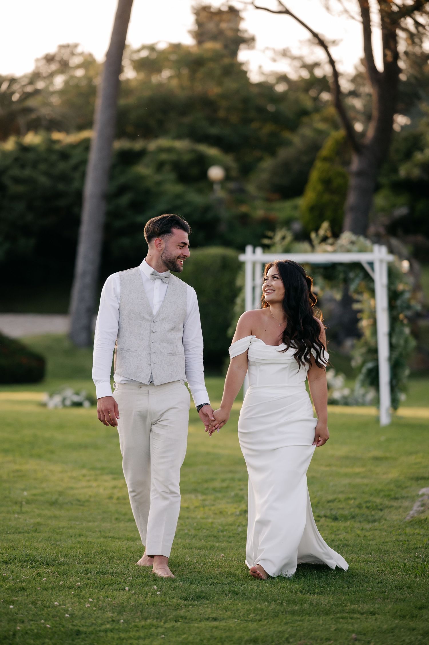 A couple in white wedding attire walks hand in hand across a grassy lawn at sunset.