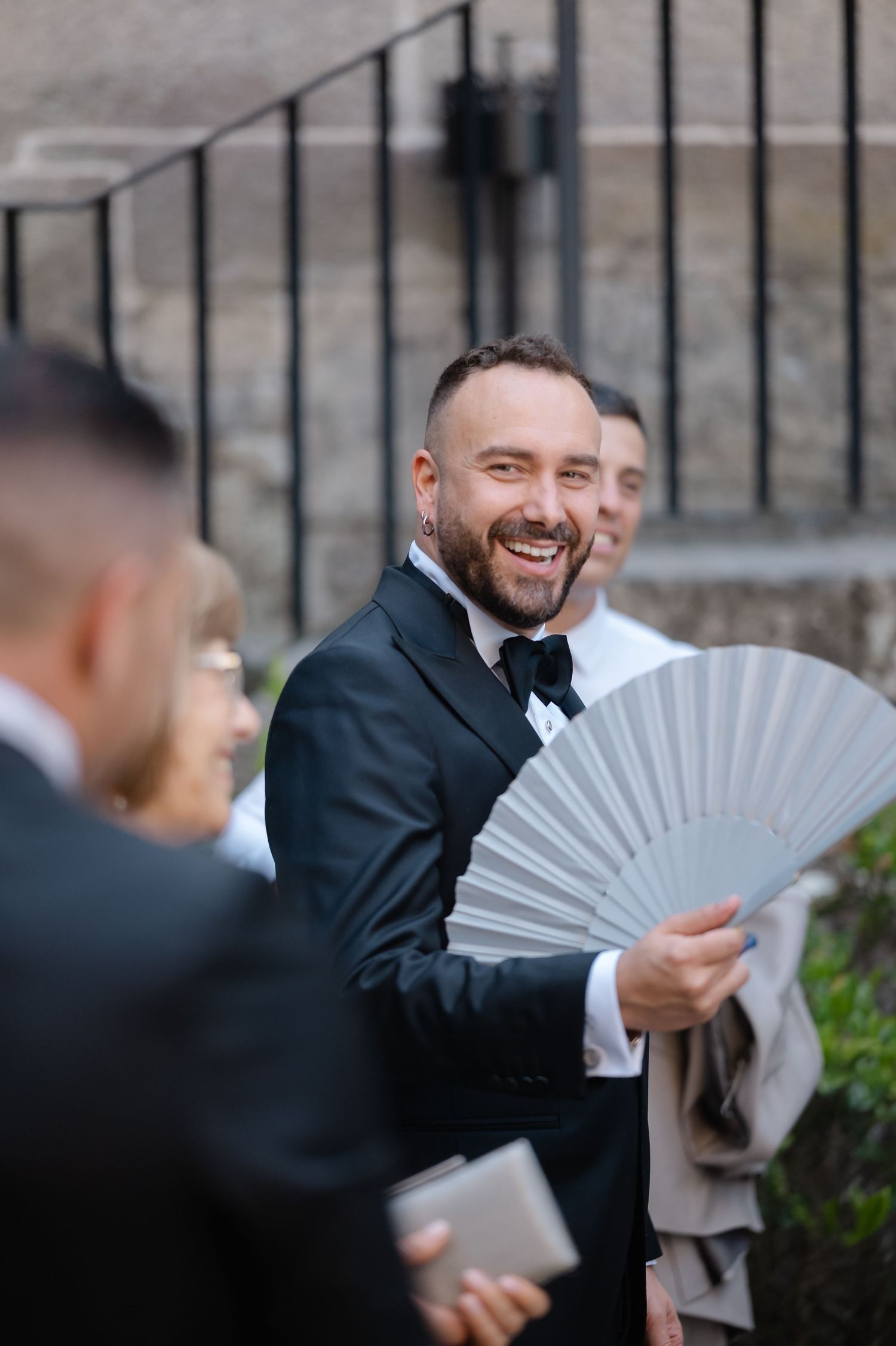 People in formal attire holding decorative paper fans while laughing and smiling outdoors.