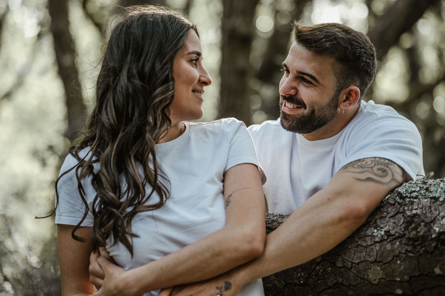 A romantic couple embraces while leaning against a tree in a dreamy outdoor forest setting.