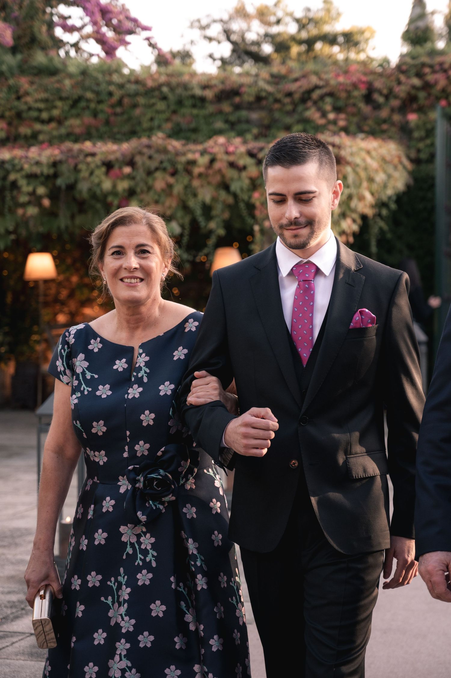 Elegant pair in formal attire walking arm-in-arm at outdoor evening event with floral backdrop and warm lighting.